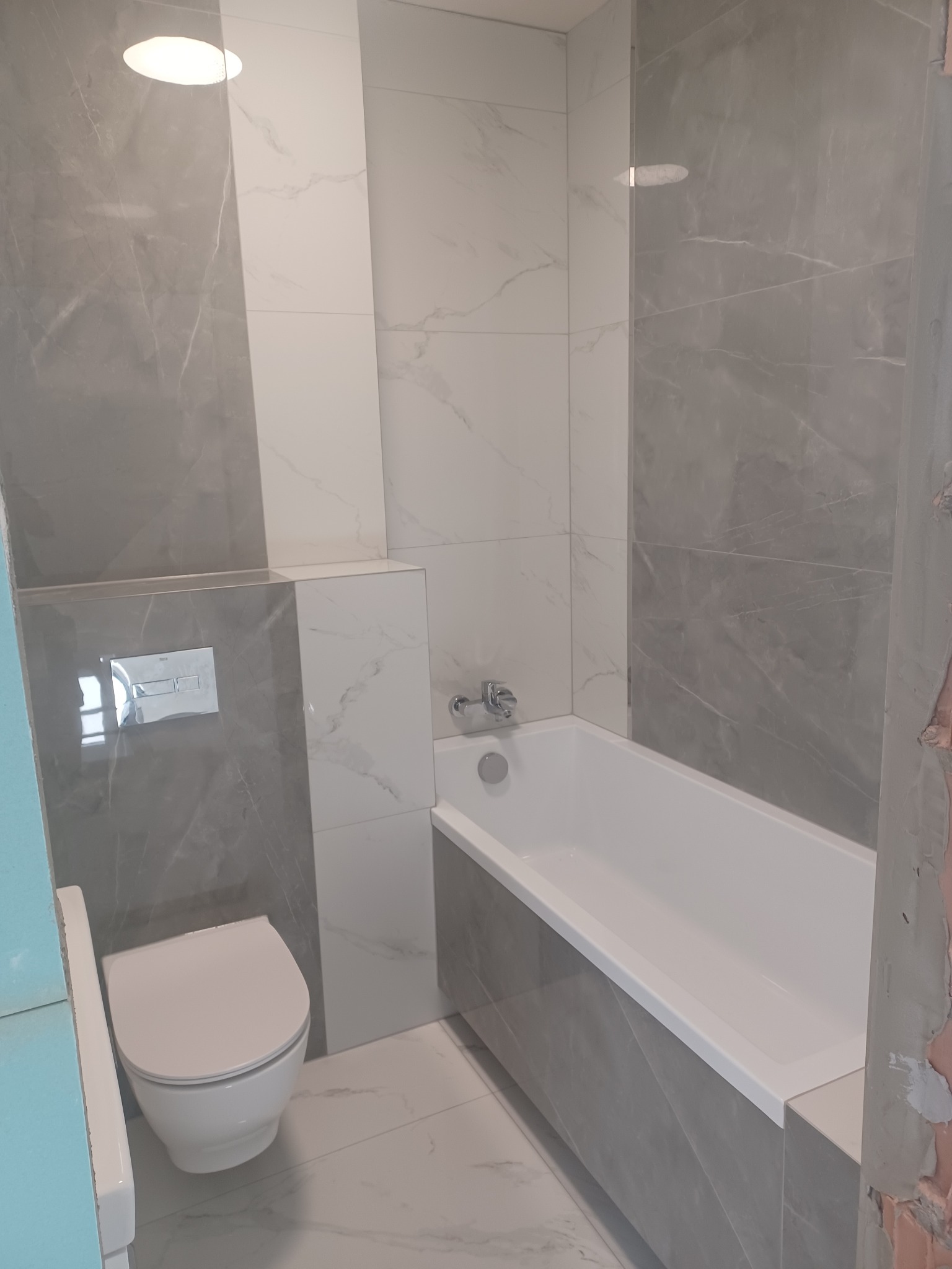 Wykończona łazienka z wanną i toaletą w szaro-białej kolorystyce. Widoczne płytki imitujące marmur na ścianach i podłodze. Nowoczesna armatura.