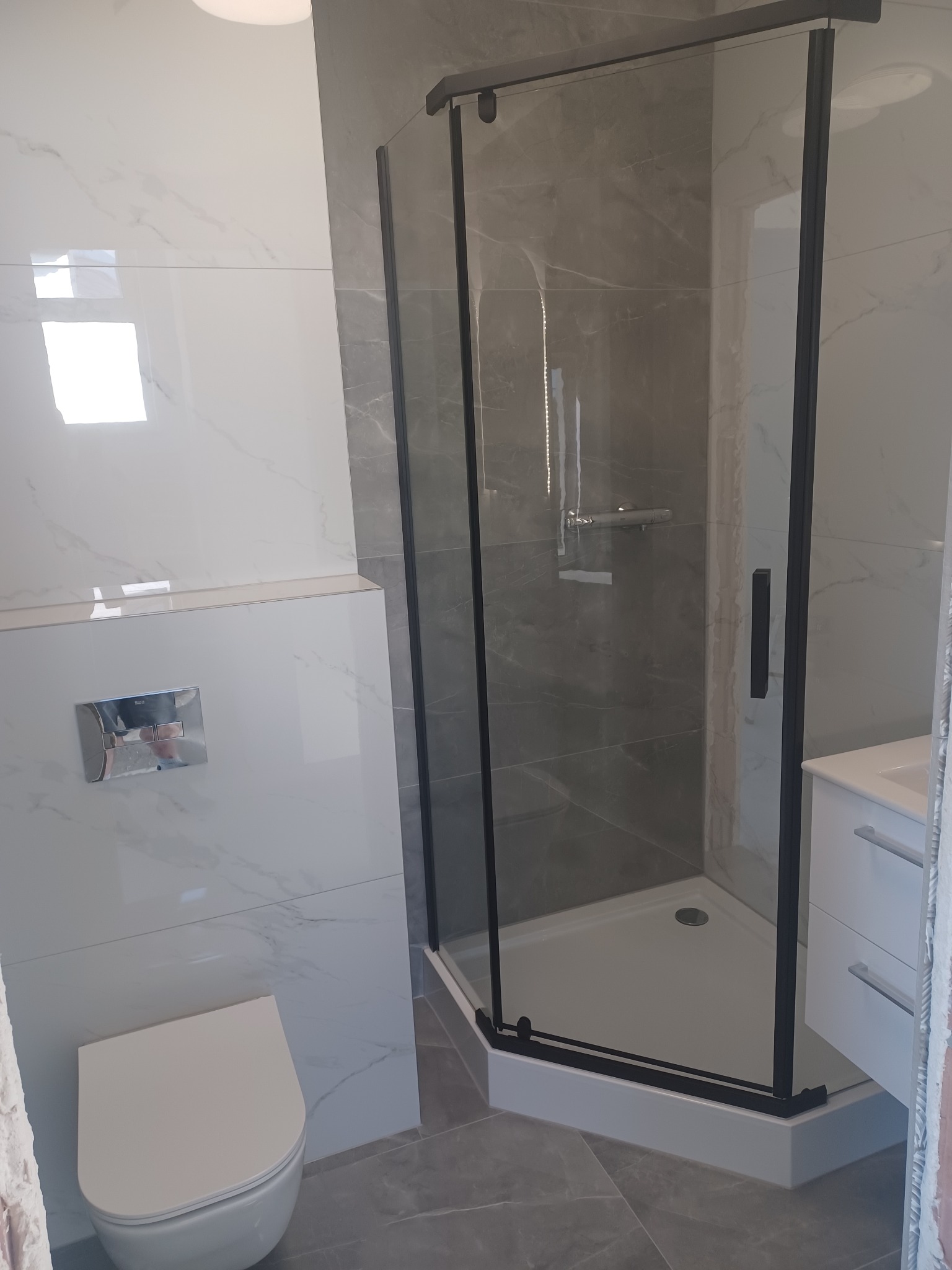 Nowoczesna łazienka z kabiną prysznicową w czarnej ramie, białą ceramiką i szarymi płytkami imitującymi marmur. Minimalistyczny design i eleganckie wykończenie.