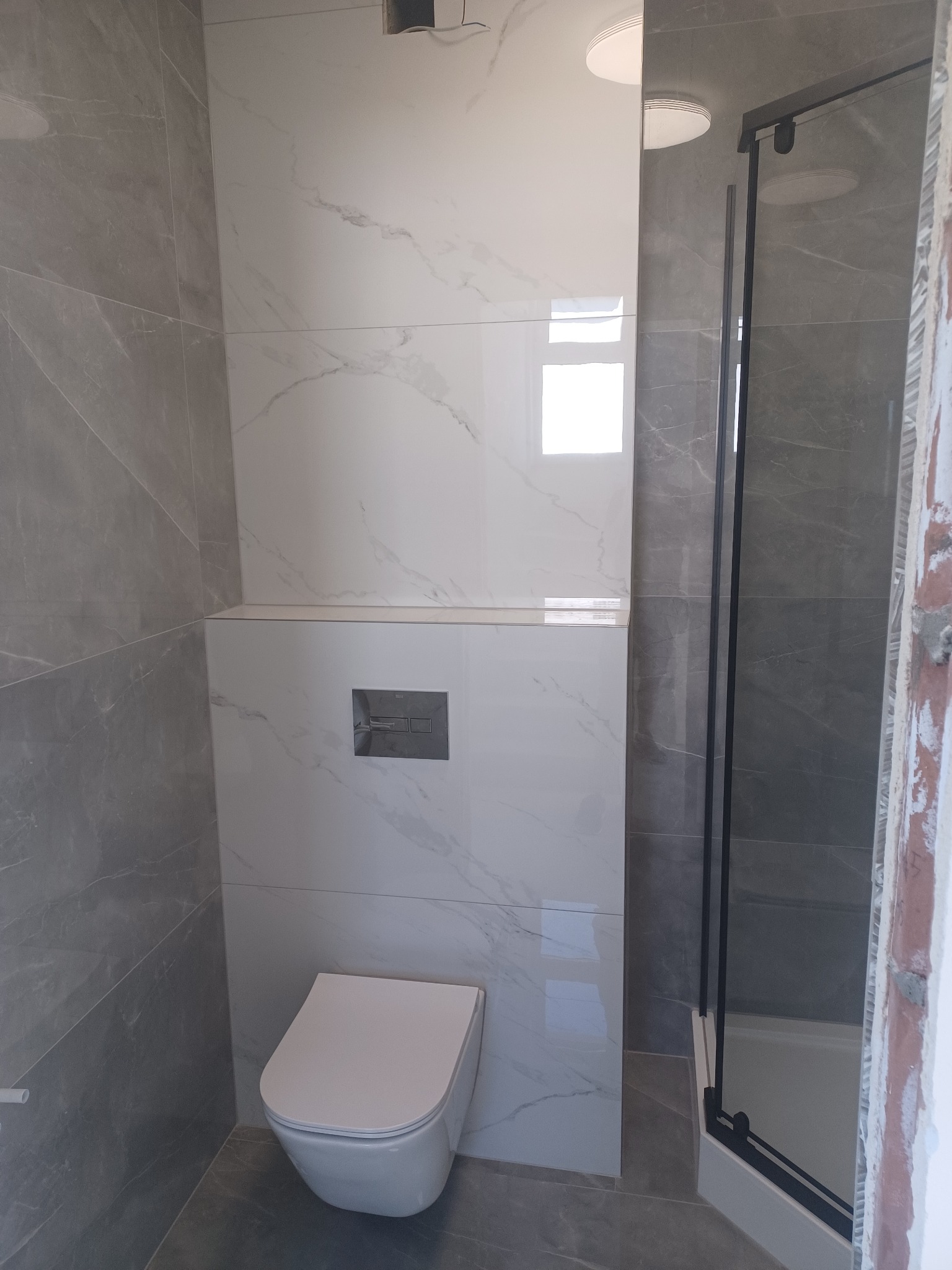 Nowoczesna łazienka z szarymi i białymi płytkami imitującymi marmur, podwieszana toaleta, czarna kabina prysznicowa i minimalistyczny design.