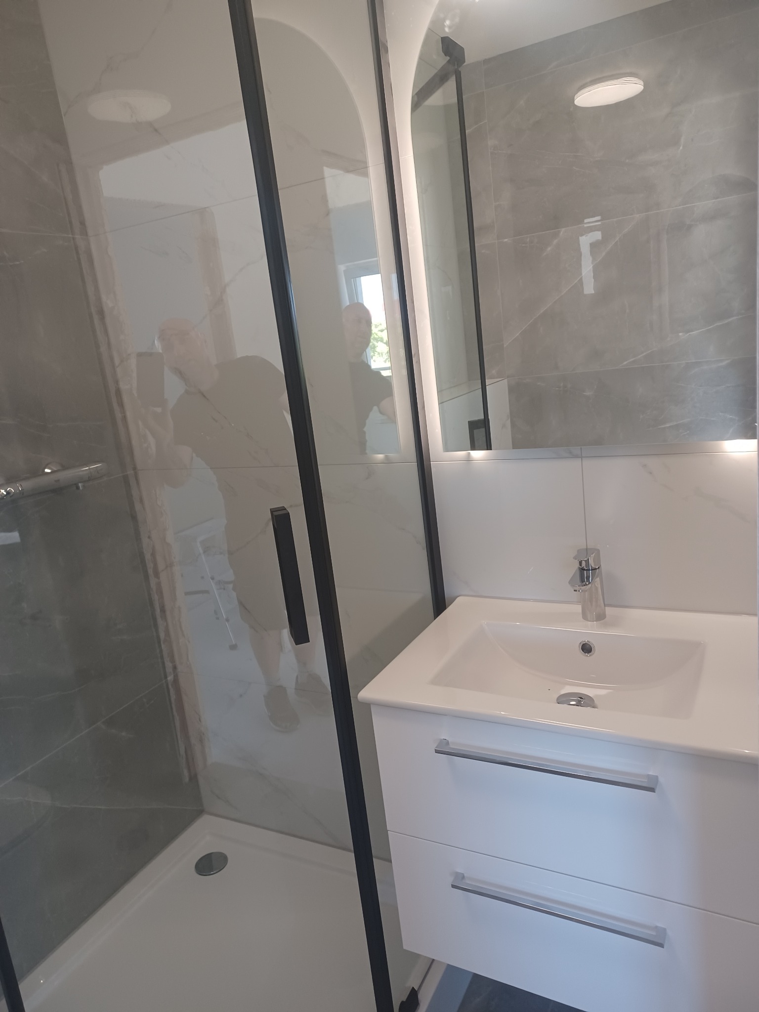 Nowoczesna łazienka po remoncie: kabina prysznicowa z czarną ramą, umywalka z szafką w bieli, szare płytki imitujące marmur, oświetlenie LED nad lustrem.