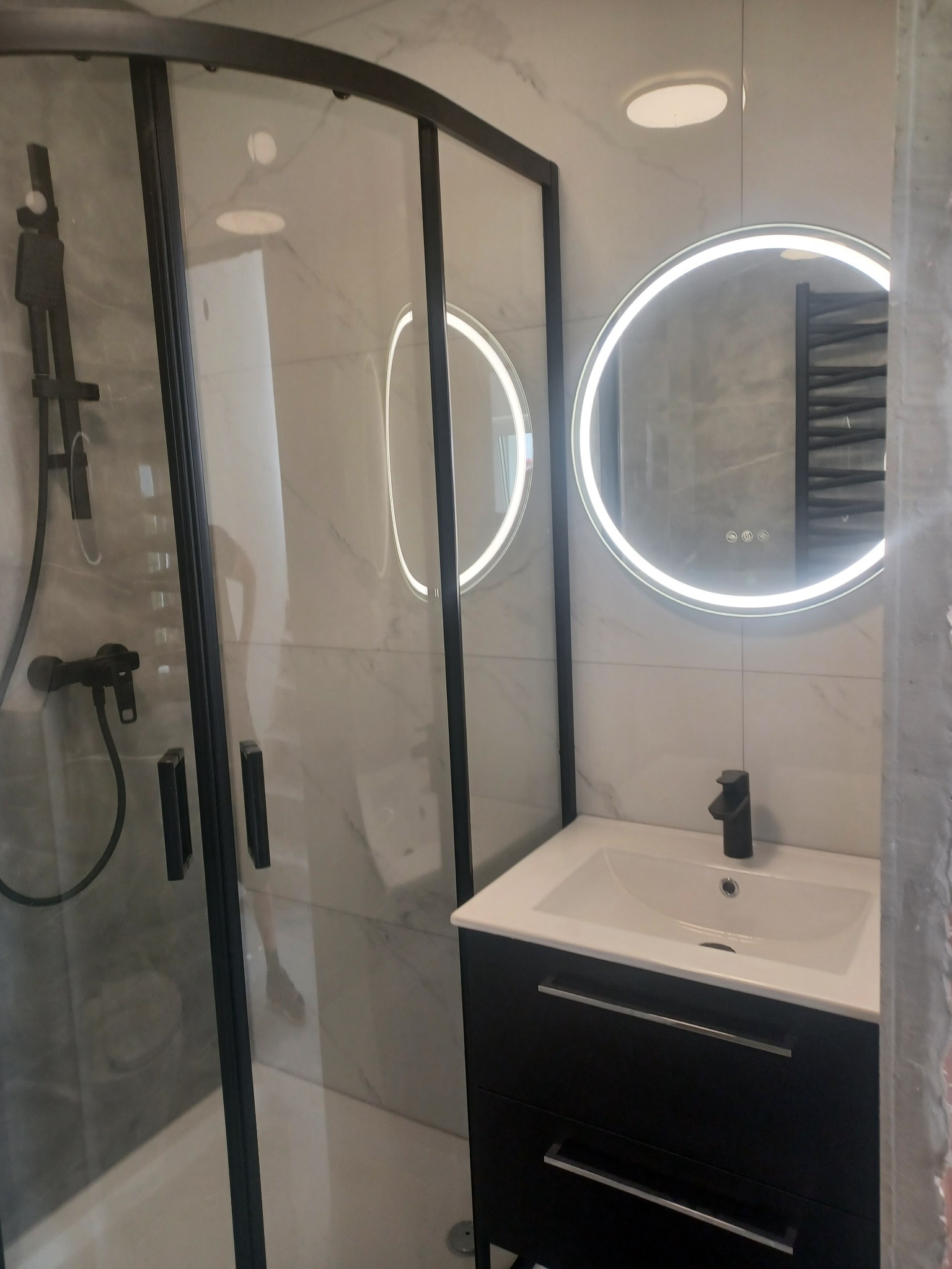 Nowoczesna łazienka z kabiną prysznicową typu walk-in, okrągłym lustrem LED i czarną armaturą. Białe płytki imitujące marmur. Minimalistyczny styl.