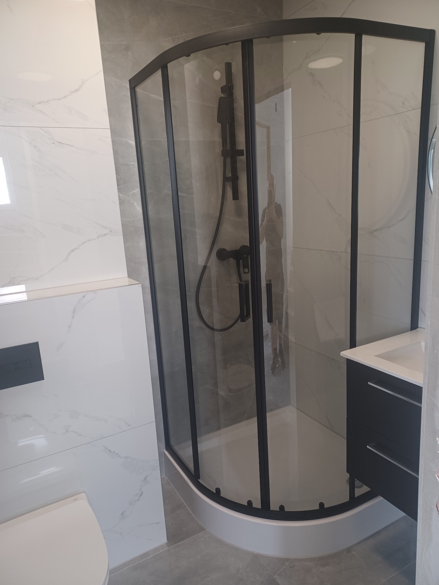 Nowoczesna kabina prysznicowa z czarnymi profilami i armaturą, marmurowe płytki na ścianach, minimalistyczna umywalka z szafką w wykończonej łazience.