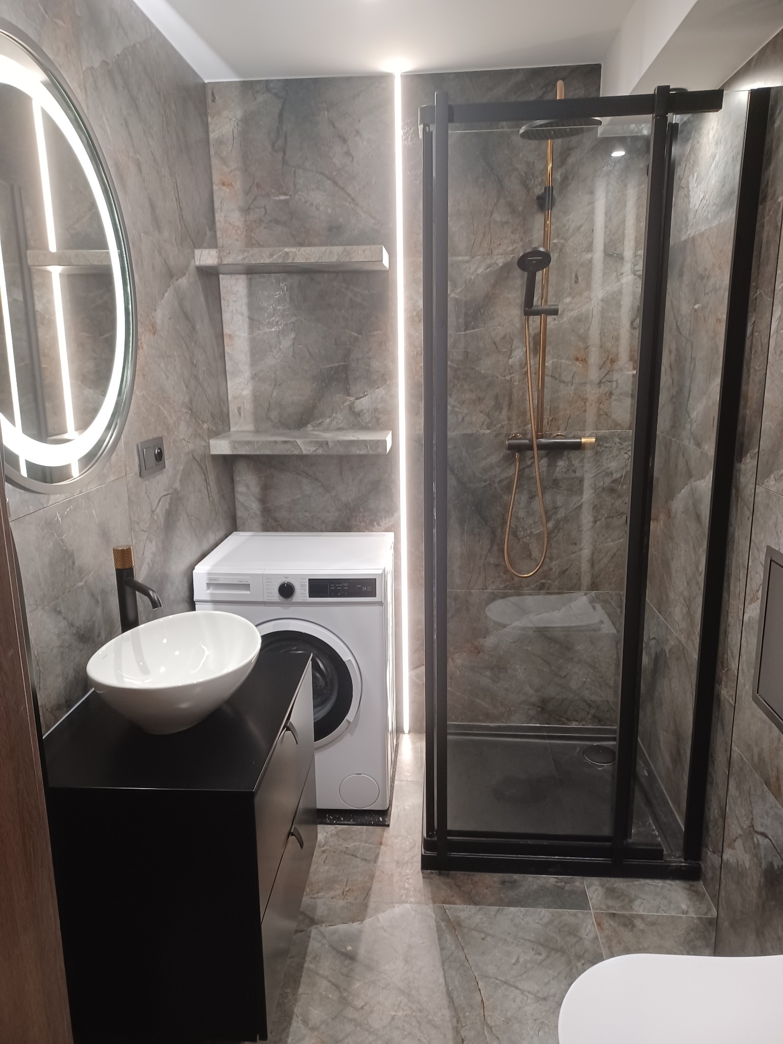 Nowoczesna łazienka z kabiną prysznicową typu walk-in, umywalką nablatową na czarnej szafce i pralką. Płytki imitujące marmur na ścianach i podłodze. Oświetlenie LED.