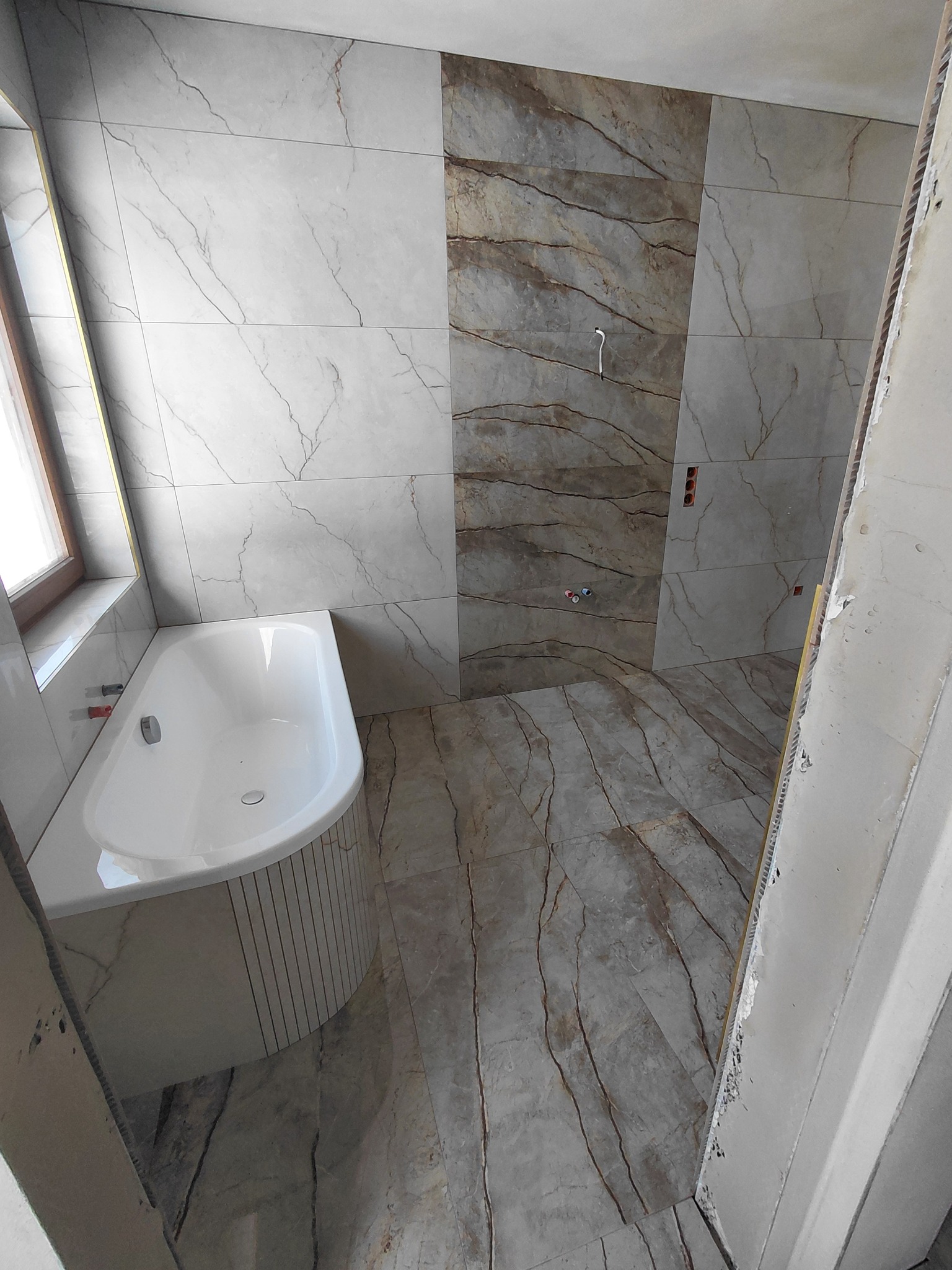 Łazienka w trakcie wykańczania: biała wanna narożna, marmurowe płytki na ścianach i podłodze z widocznymi instalacjami. Częściowo wykończone wnętrze.