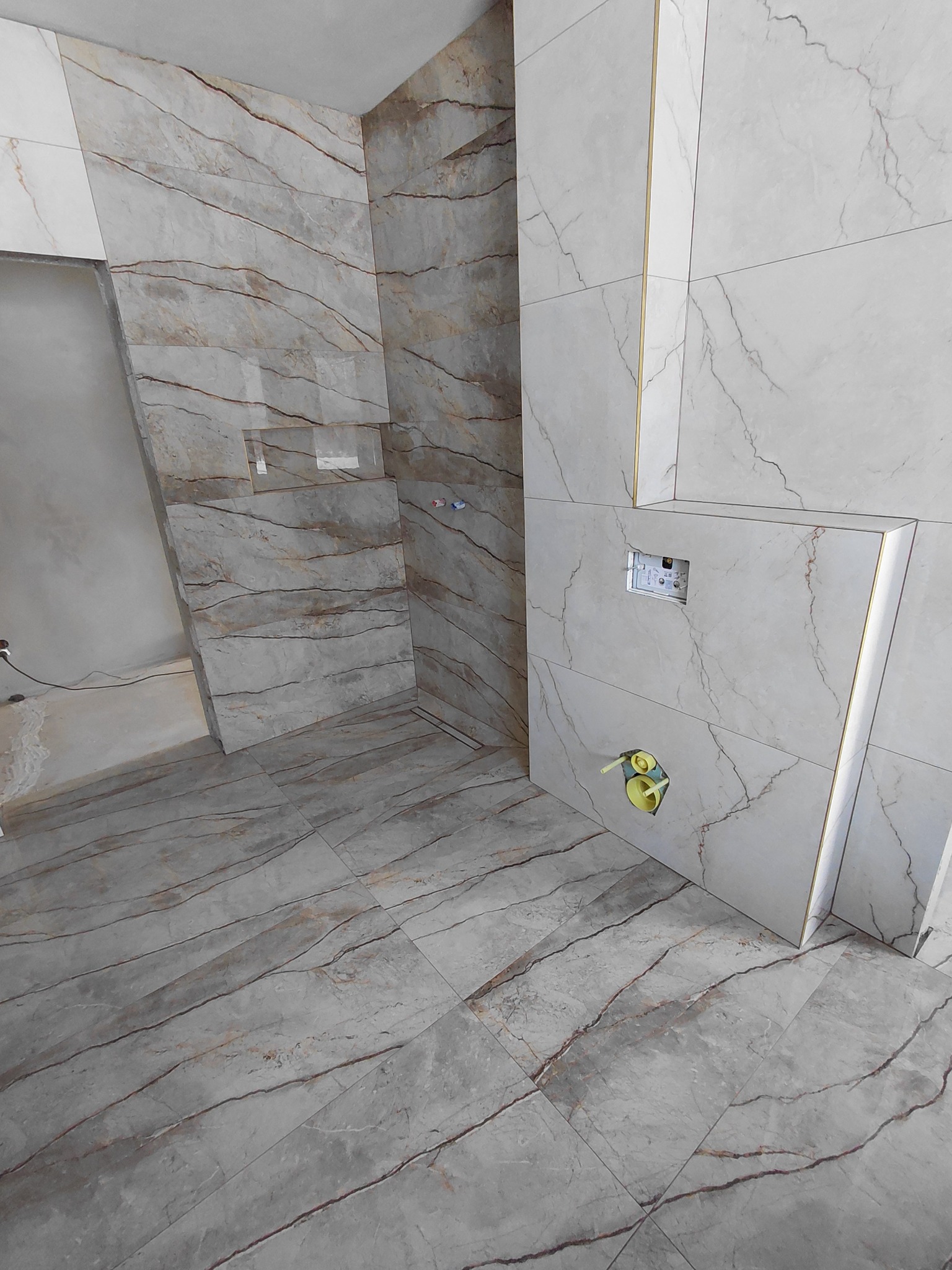 Wykończona łazienka z marmurowymi płytkami na ścianach i podłodze. Widoczne elementy instalacji sanitarnej i stelaż podtynkowy WC. Estetyczne wykończenie z dbałością o detale.