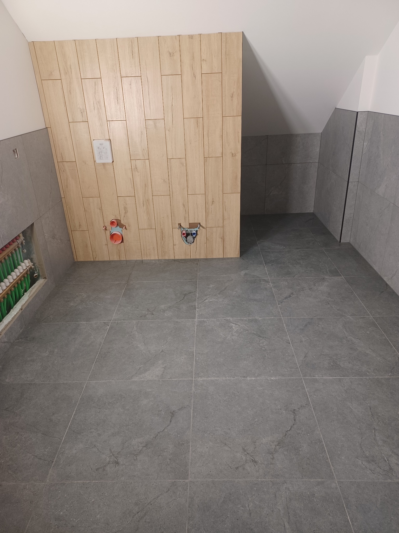 Wykończona łazienka w Woli Zgłobieńskiej: szare płytki podłogowe i ścienne, drewnopodobna zabudowa z instalacją. Widok na wnętrze w trakcie prac wykończeniowych.