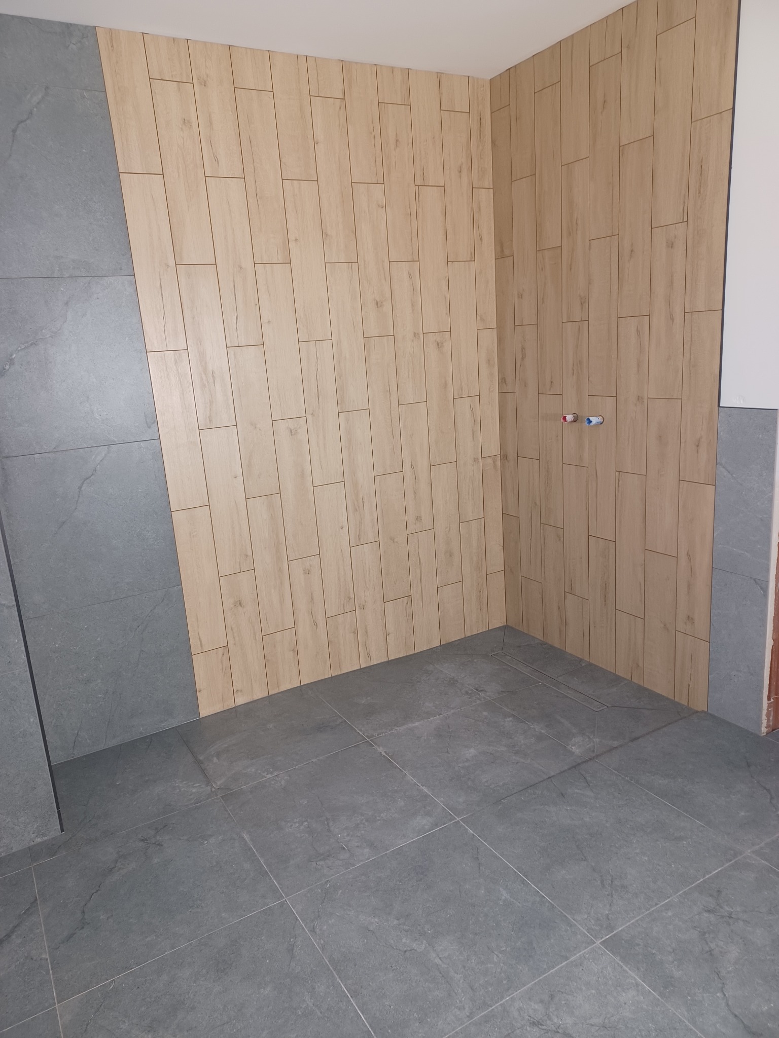 Wykończona łazienka z szarą podłogą i ścianą z płytek imitujących drewno. Widoczne przyłącza wodne. Ukończony remont łazienki w nowoczesnym stylu.