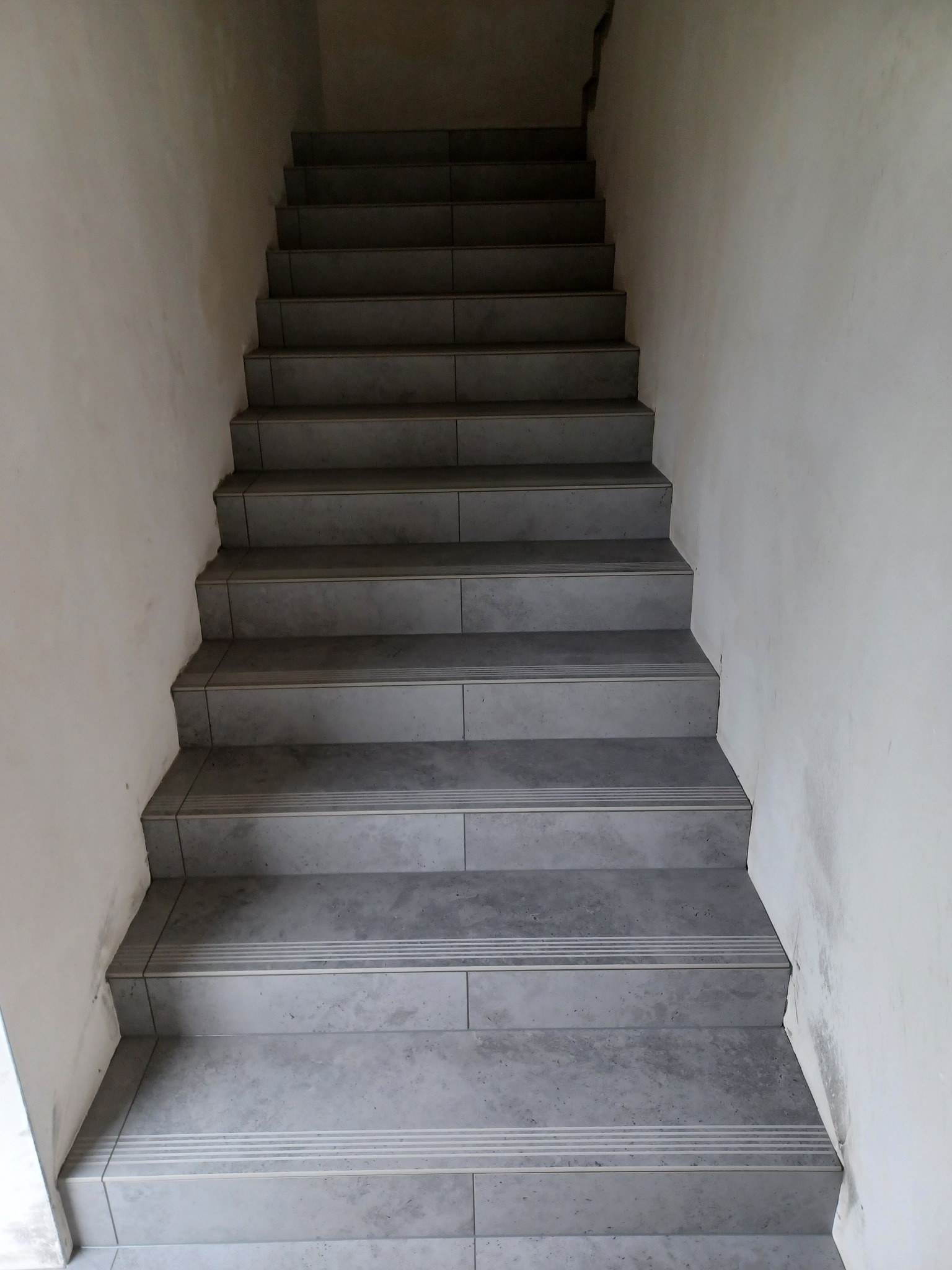 Schody wyłożone szarą, matową płytką ceramiczną z delikatnym wzorem, prowadzące w górę. Widoczne białe ściany klatki schodowej. Ujęcie z dołu, podkreślające pionowy układ.