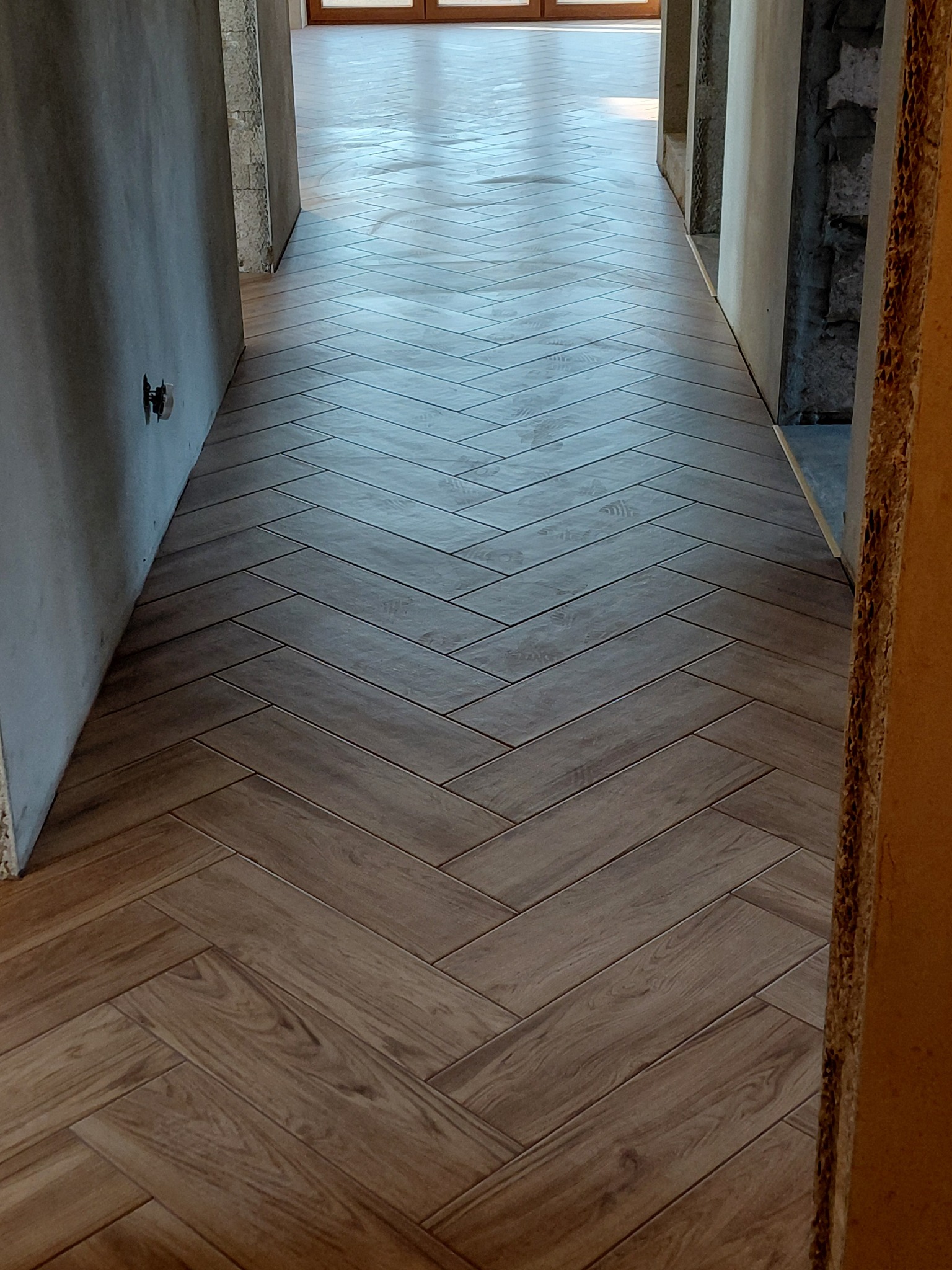 Ułożona w jodełkę, brązowa podłoga z płytek imitujących drewno w korytarzu. Widoczne surowe ściany i fragment futryny. Nowoczesny design.