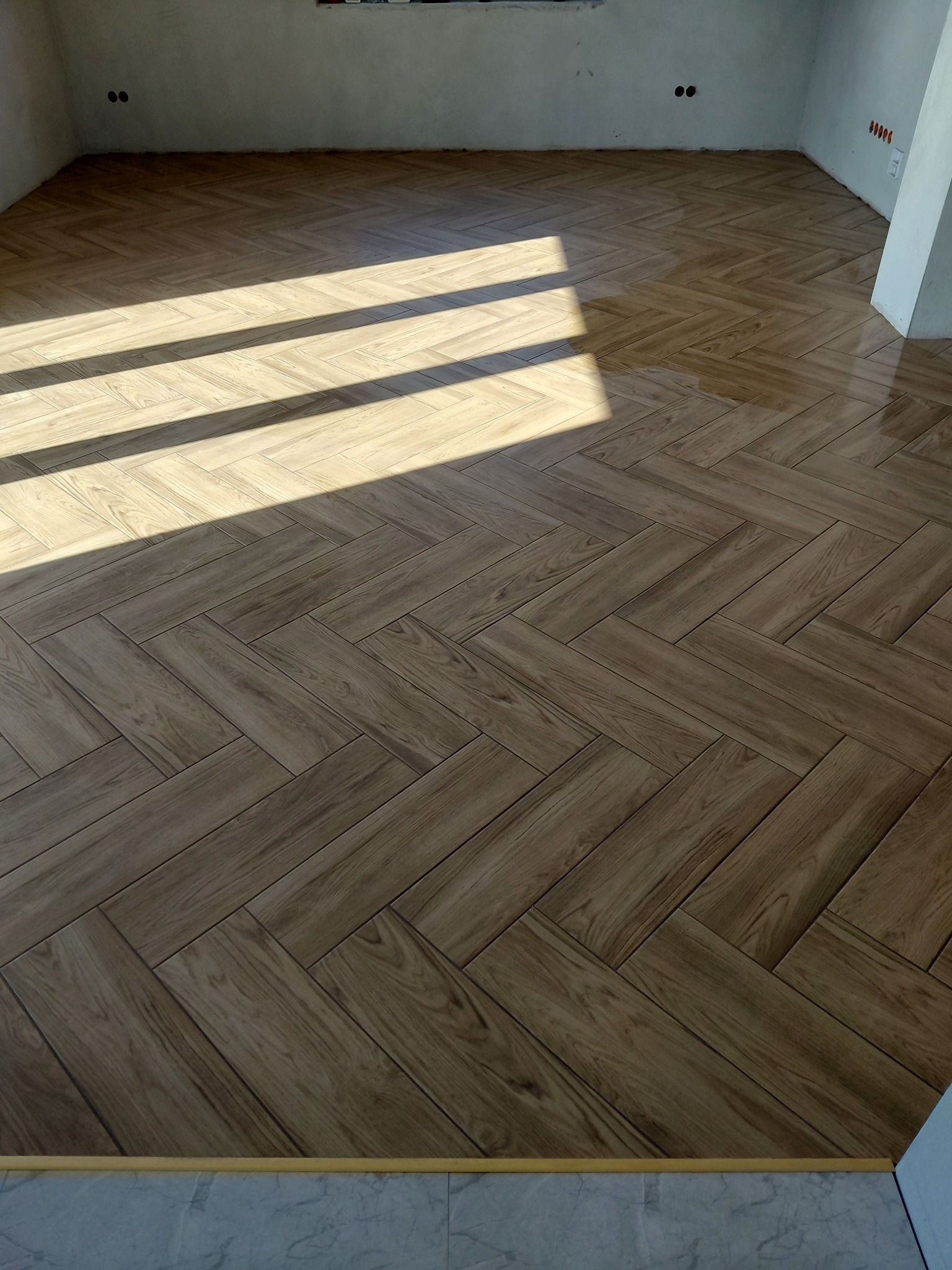 Ukończona podłoga z płytek imitujących drewno, ułożonych w jodełkę, z widocznymi promieniami słońca. Ściany w stanie surowym, widoczne gniazdka elektryczne.