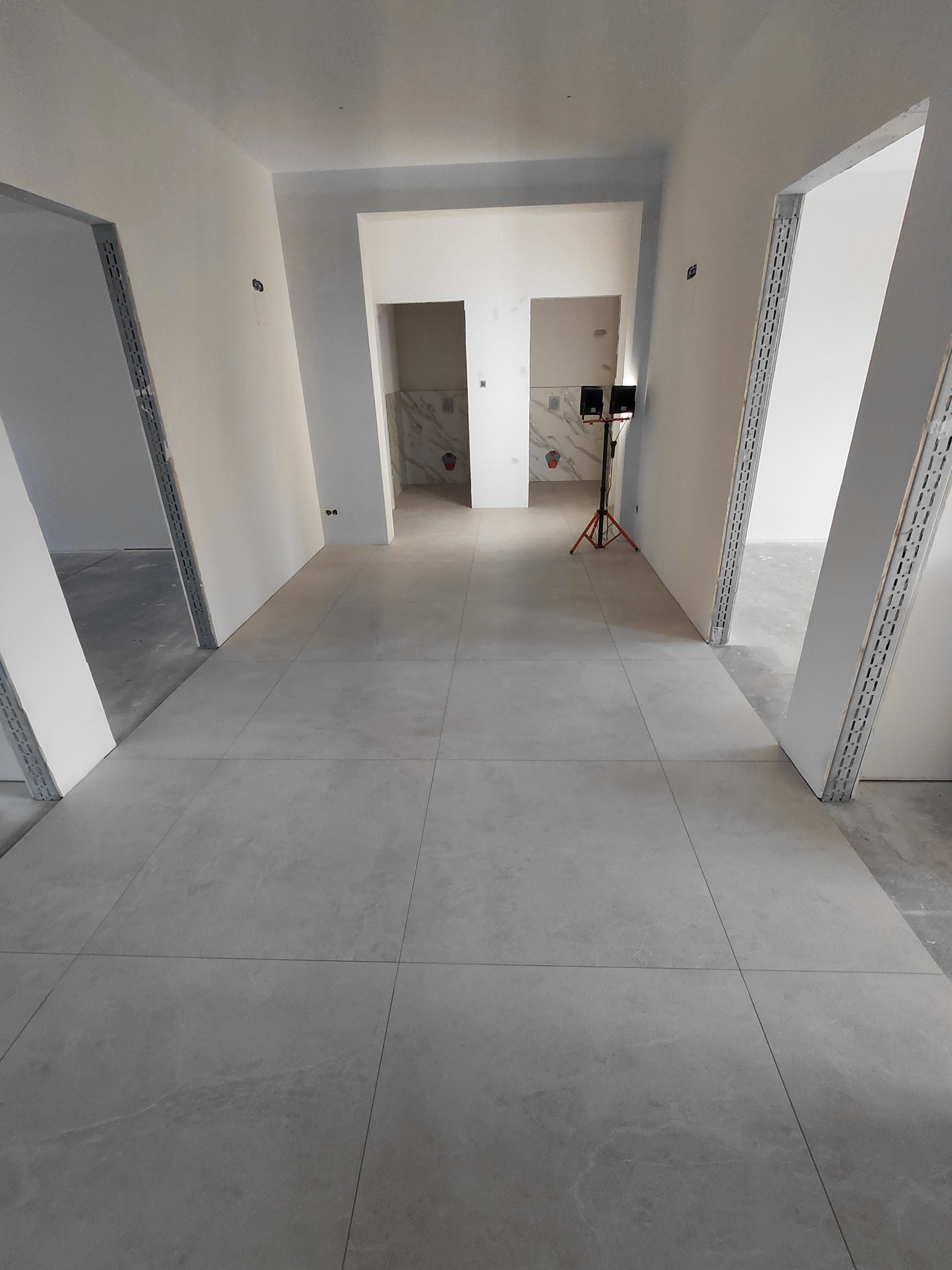 Ułożona szara podłoga z płytek w korytarzu, widoczne przygotowane ościeżnice drzwiowe i fragment wykończonej łazienki w głębi. Na podłodze stoi lampa budowlana.