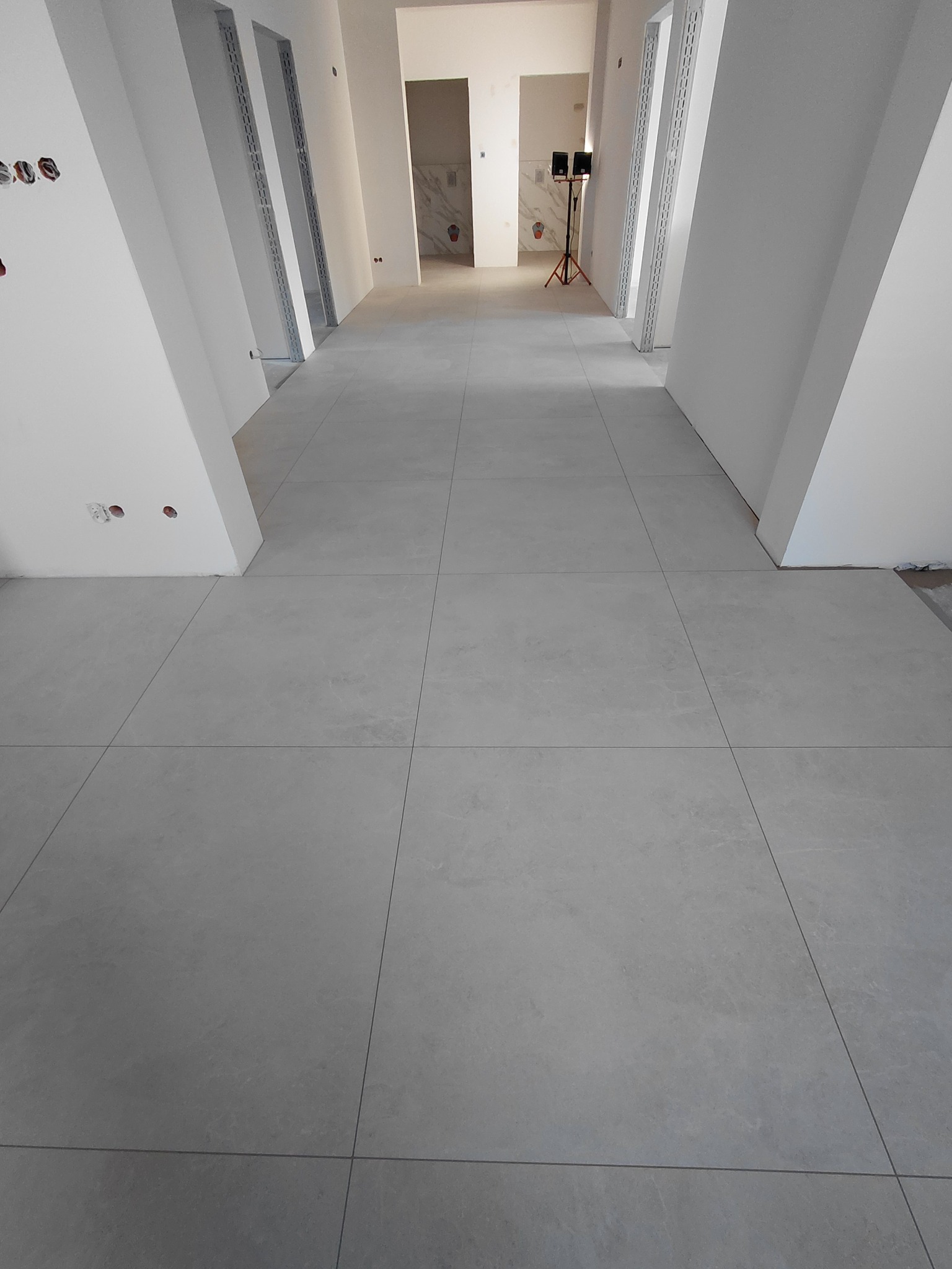 Jasnoszara, duża płytka podłogowa w korytarzu z białymi ścianami i otworami drzwiowymi. W tle widoczne pomieszczenia w trakcie wykańczania.