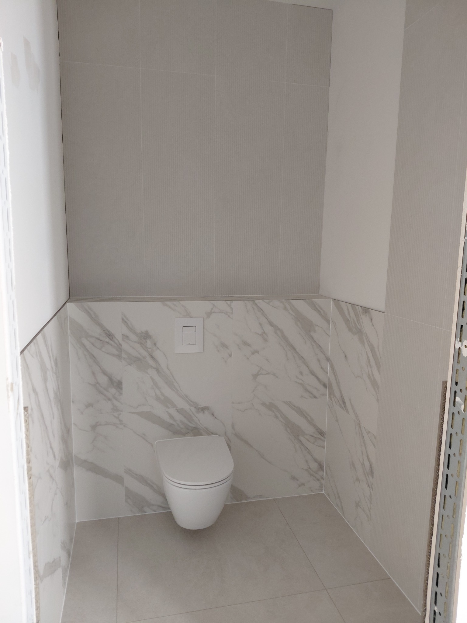 Wykończona łazienka z toaletą wiszącą i płytkami imitującymi marmur na ścianach do połowy wysokości. Górna część ścian wyłożona szarymi, podłużnymi płytkami.