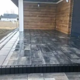 Wyłożony kostką brukową taras z ciemnoszarych płyt, z ciemną listwą krawędziową, przed budynkiem z drewnianą elewacją i roletami.