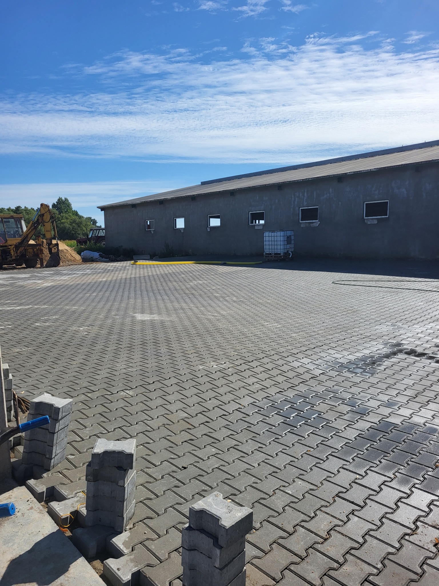 Rozległy plac wyłożony szarą kostką brukową typu behaton, z widocznym żółtym koparko-ładowarką w tle i stosem kostki na pierwszym planie, częściowo ułożonej, pod błękitnym niebem z chmurami.