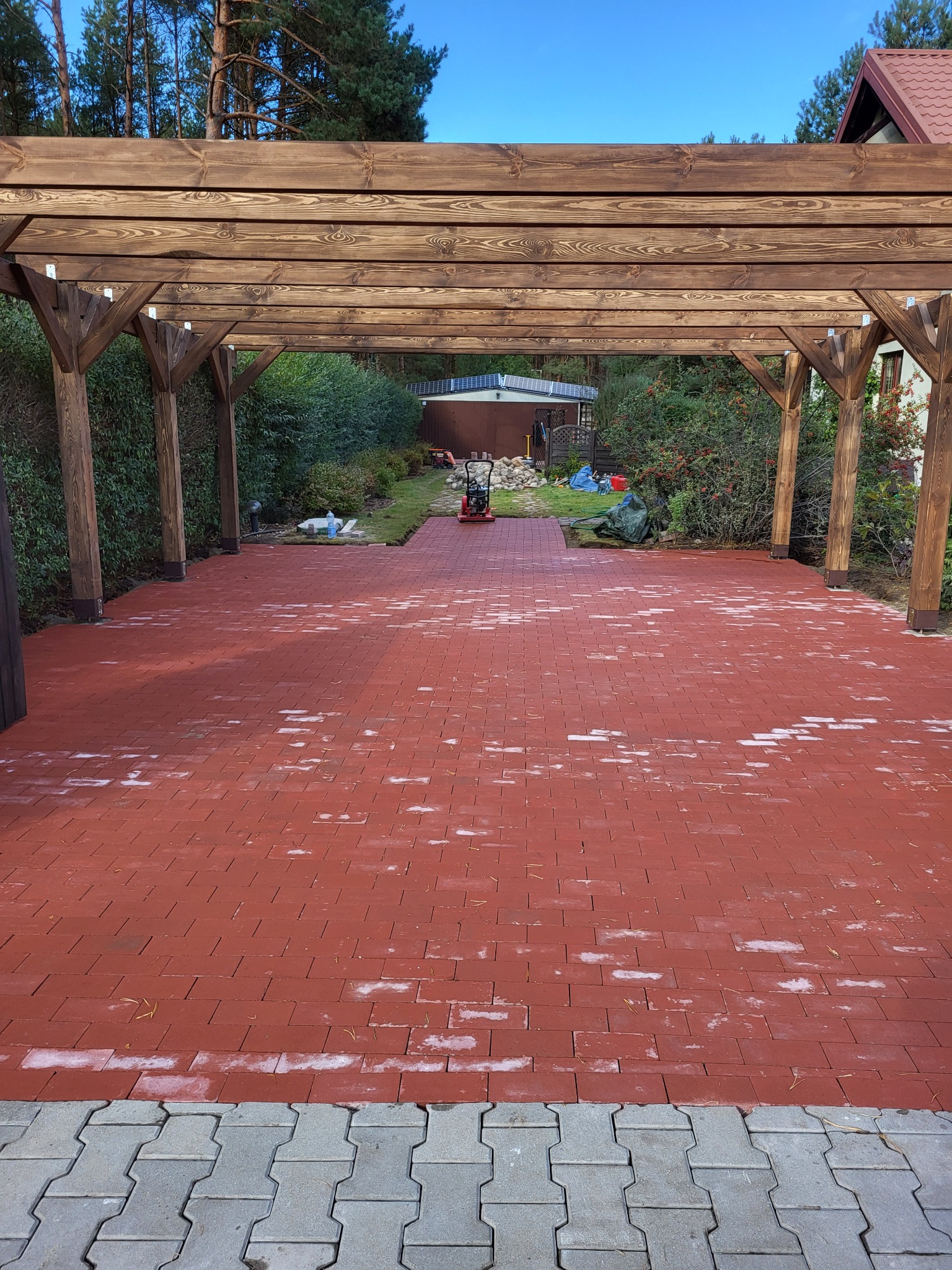 Czerwona kostka brukowa ułożona pod drewnianą pergolą, widoczne ślady fugowania, w tle trawnik i budynek gospodarczy.