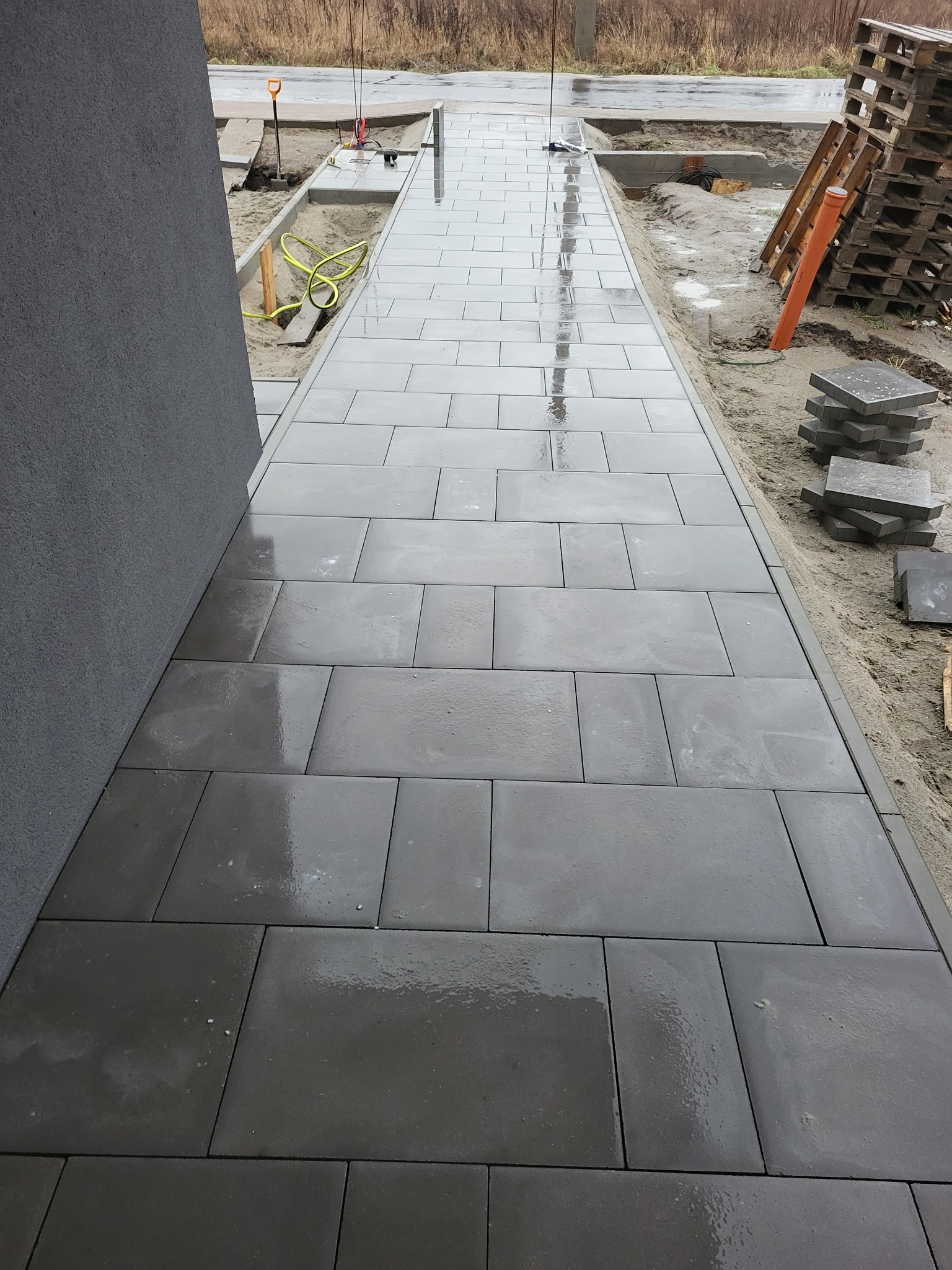 Świeżo ułożona, mokra ścieżka z szarych, prostokątnych płyt betonowych prowadząca wzdłuż ściany budynku, widoczne przygotowane podłoże i palety z kostką w tle.