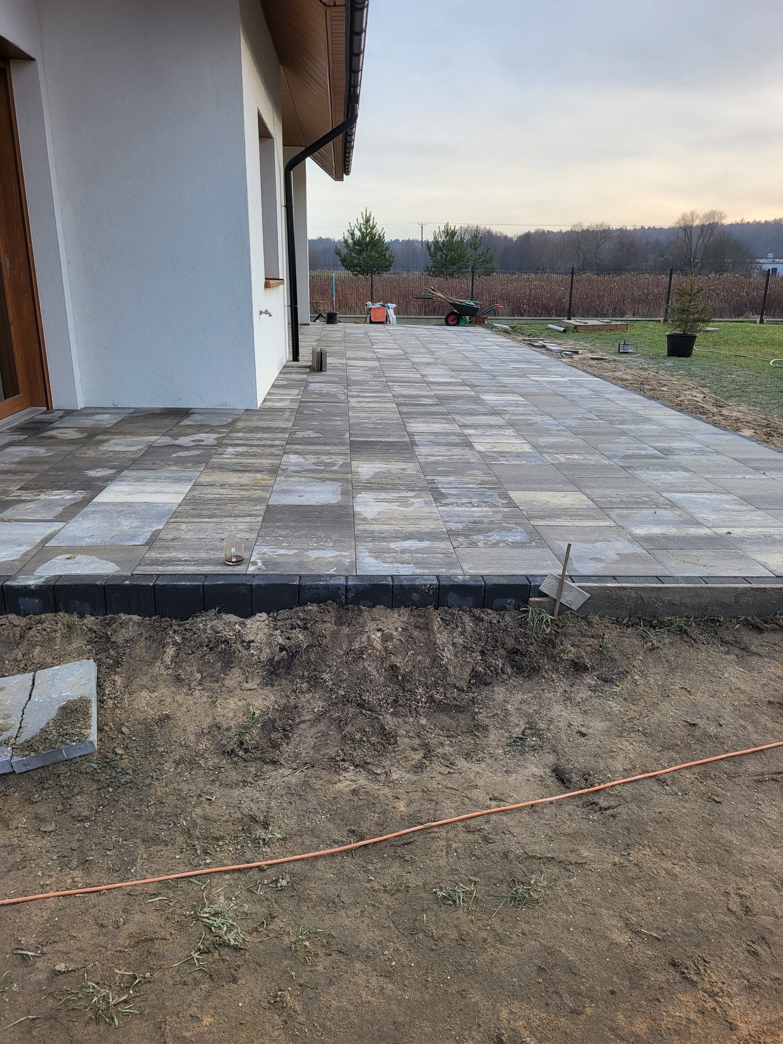 Fragment posesji z nowo ułożoną kostką brukową w odcieniach szarości i beżu, obrzeża z czarnej kostki, ziemia przygotowana do dalszych prac wykończeniowych, w tle dom jednorodzinny i drzewa.