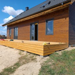 Usługi ogólnobudowlane Krzysztof Cymerys - Nowoczesny dom z drewnianą elewacją i tarasem w Szczytnie, widoczne narzędzia pracy na tarasie, błękitne niebo z chmurami w tle.