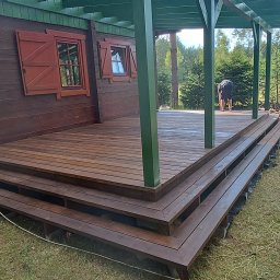 Usługi ogólnobudowlane Krzysztof Cymerys - Montaż drewnianego tarasu z trzema stopniami przy brązowym domu z zielonymi elementami, widoczne narzędzia i pracownicy w trakcie pracy.