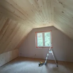 Poddasze w trakcie wykańczania, widoczne drewniane panele na skośnym suficie, ściany z płyt gipsowo-kartonowych, okno, drabina i narzędzia budowlane na podłodze.