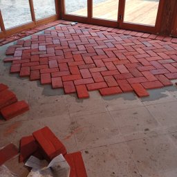 Usługi ogólnobudowlane Krzysztof Cymerys - Układanie czerwonej cegły klinkierowej na podłodze w przeszklonym pomieszczeniu. Widoczne częściowo ułożone cegły i zapas materiału obok. Praca w toku.
