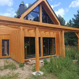 Usługi ogólnobudowlane Krzysztof Cymerys - Nowoczesny dom z jasnego drewna z dużymi, trójkątnymi oknami w szczycie i dobudowaną drewnianą konstrukcją pergoli. Czarne ramy okien i komin, otoczenie leśne.