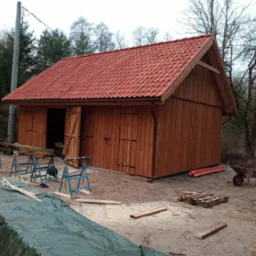 Nowo wybudowany, drewniany budynek gospodarczy z czerwoną dachówką, częściowo otwartymi drzwiami, widoczne narzędzia i materiały budowlane na placu budowy.