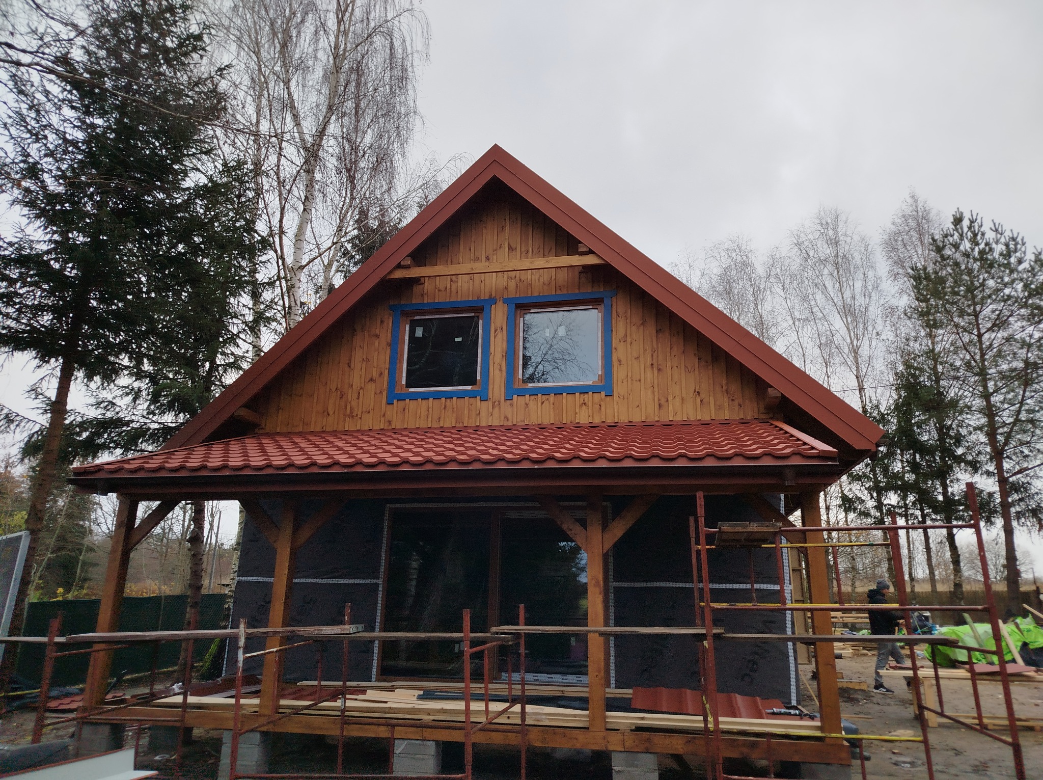 Surowy stan budowy domu z drewnianą elewacją, czerwonym dachem i niebieskimi obramowaniami okiennymi, otoczonego rusztowaniami i drzewami.