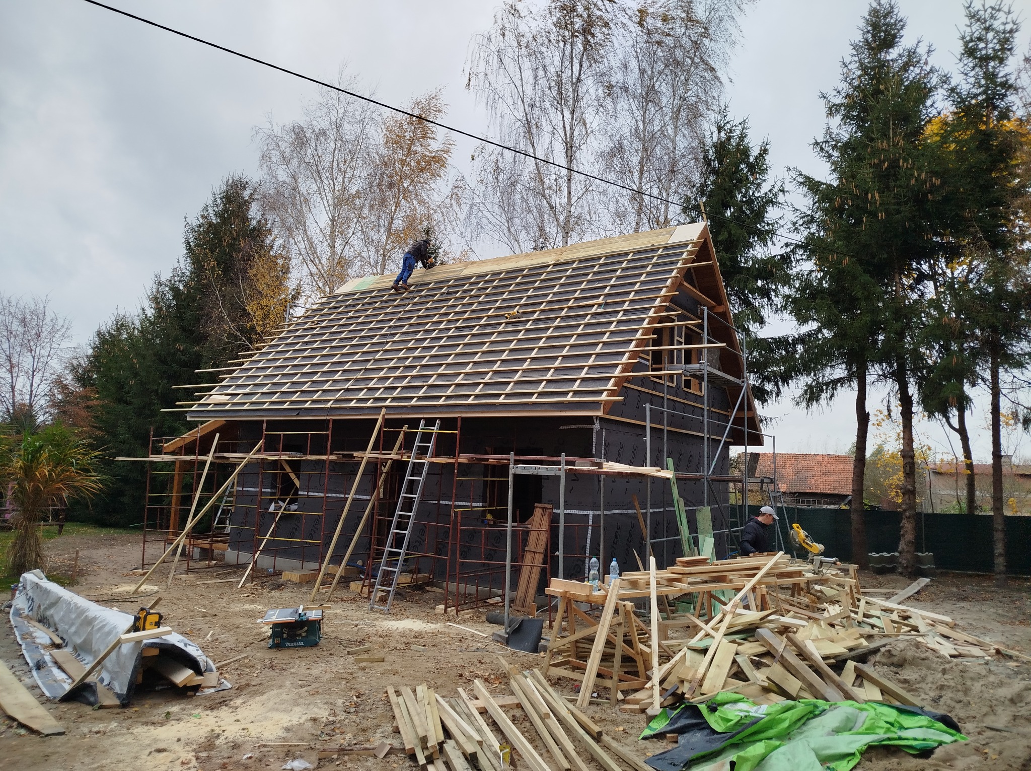 Budowa domu w technologii szkieletowej: montaż więźby dachowej, widoczne rusztowania, izolacja ścian zewnętrznych czarną membraną, pracownicy na dachu i przy pile, plac budowy z materiałami.