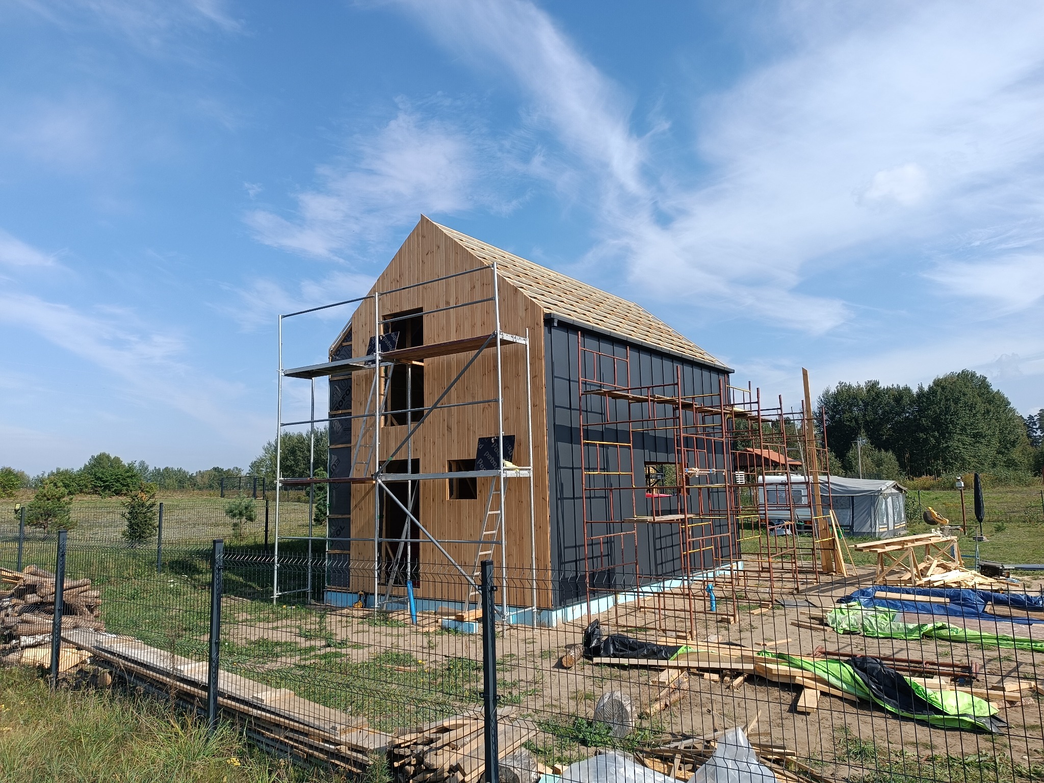 Budowa domu w technologii szkieletowej w trakcie realizacji, widoczne rusztowania, deski elewacyjne i niebieskie zabezpieczenia fundamentów, błękitne niebo w tle.