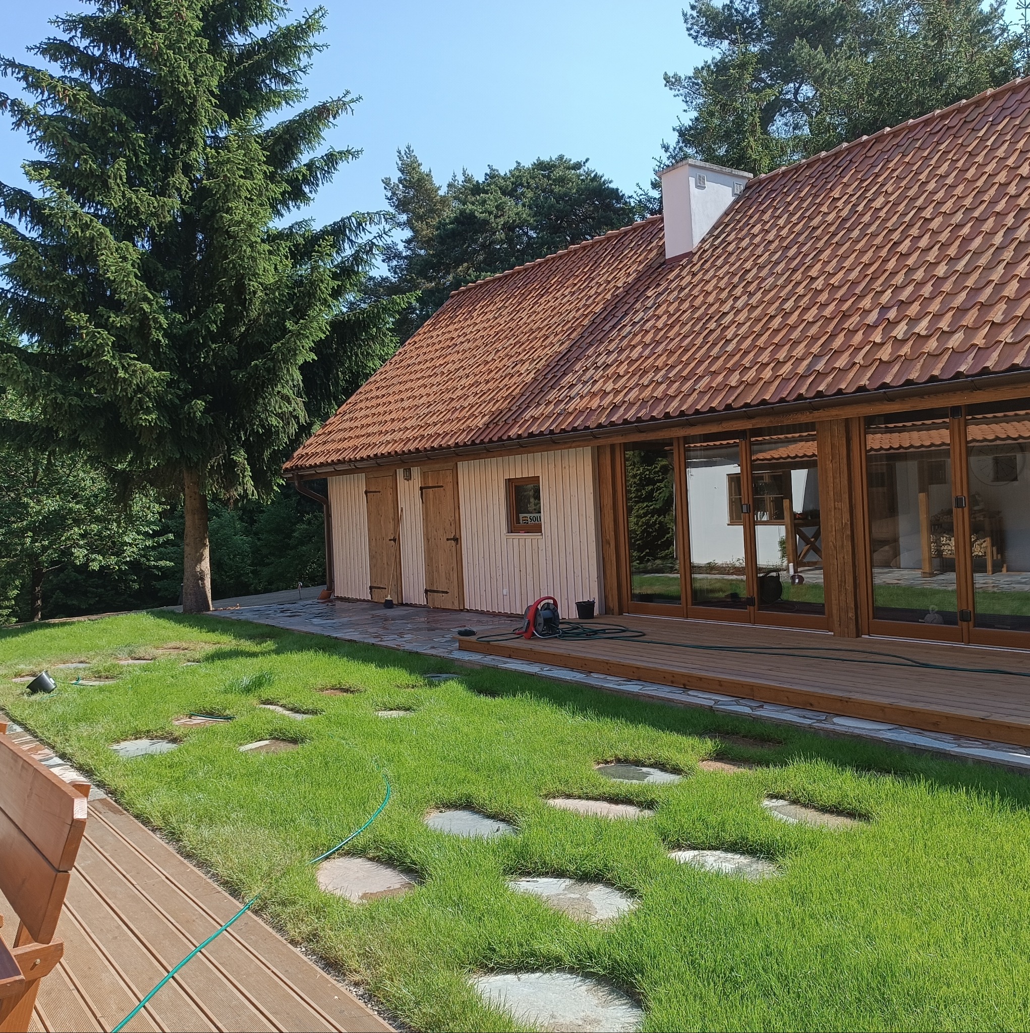 Nowoczesny dom z czerwonym dachem, drewnianą elewacją i dużymi oknami, otoczony zadbanym trawnikiem z kamiennymi ścieżkami i drewnianym tarasem.