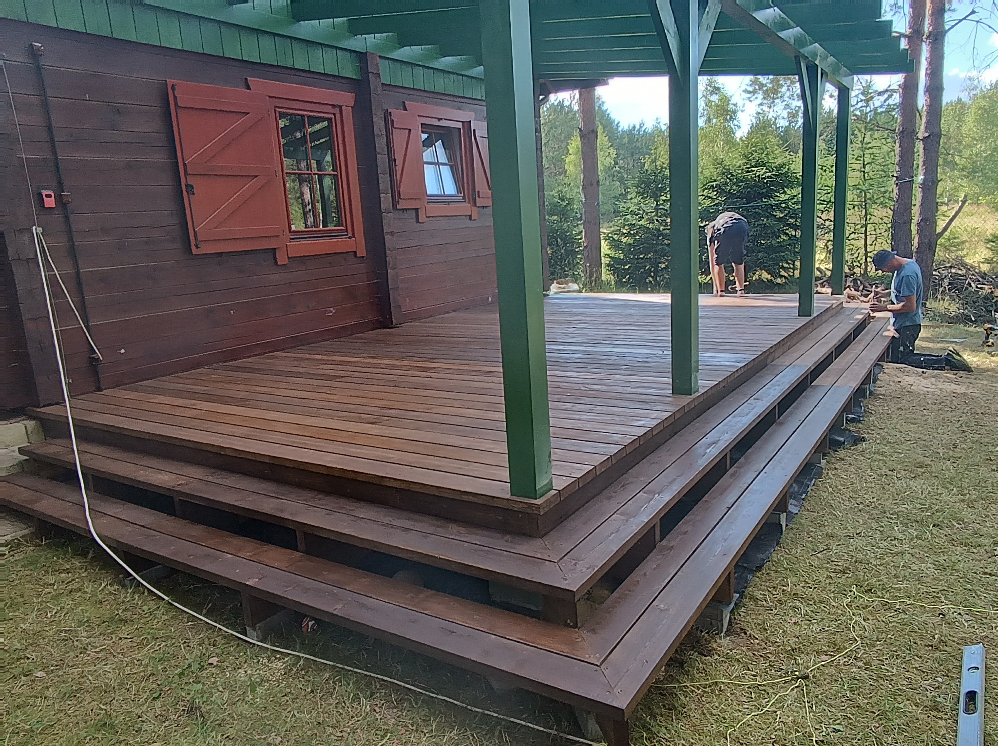 Montaż drewnianego tarasu z trzema stopniami przy brązowym domu z zielonymi elementami, widoczne narzędzia i pracownicy w trakcie pracy.