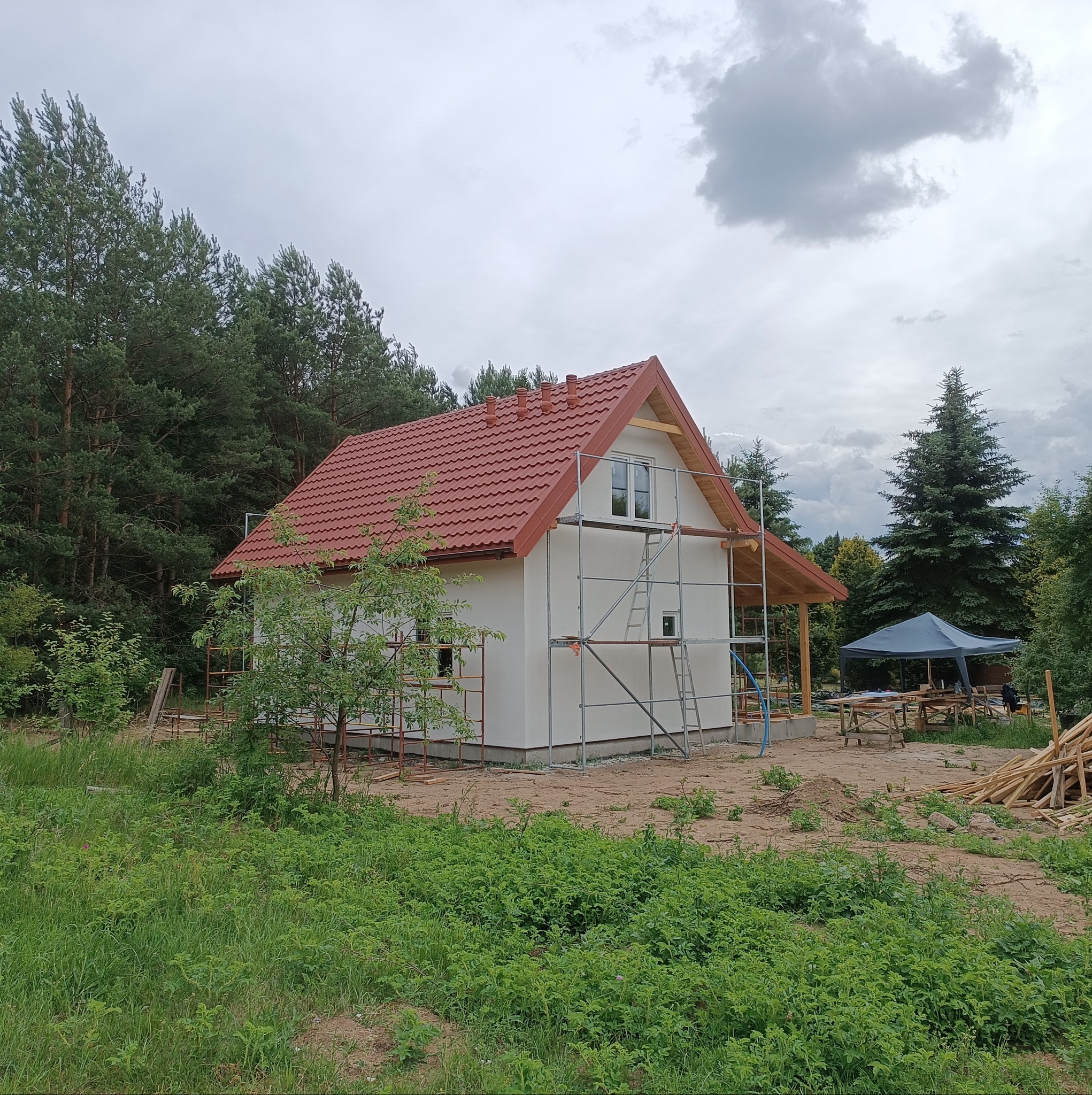 Surowy, biały budynek z czerwoną dachówką, otoczony rusztowaniami, na tle lasu i pochmurnego nieba. Widoczne drewniane elementy konstrukcyjne i stos drewna obok.