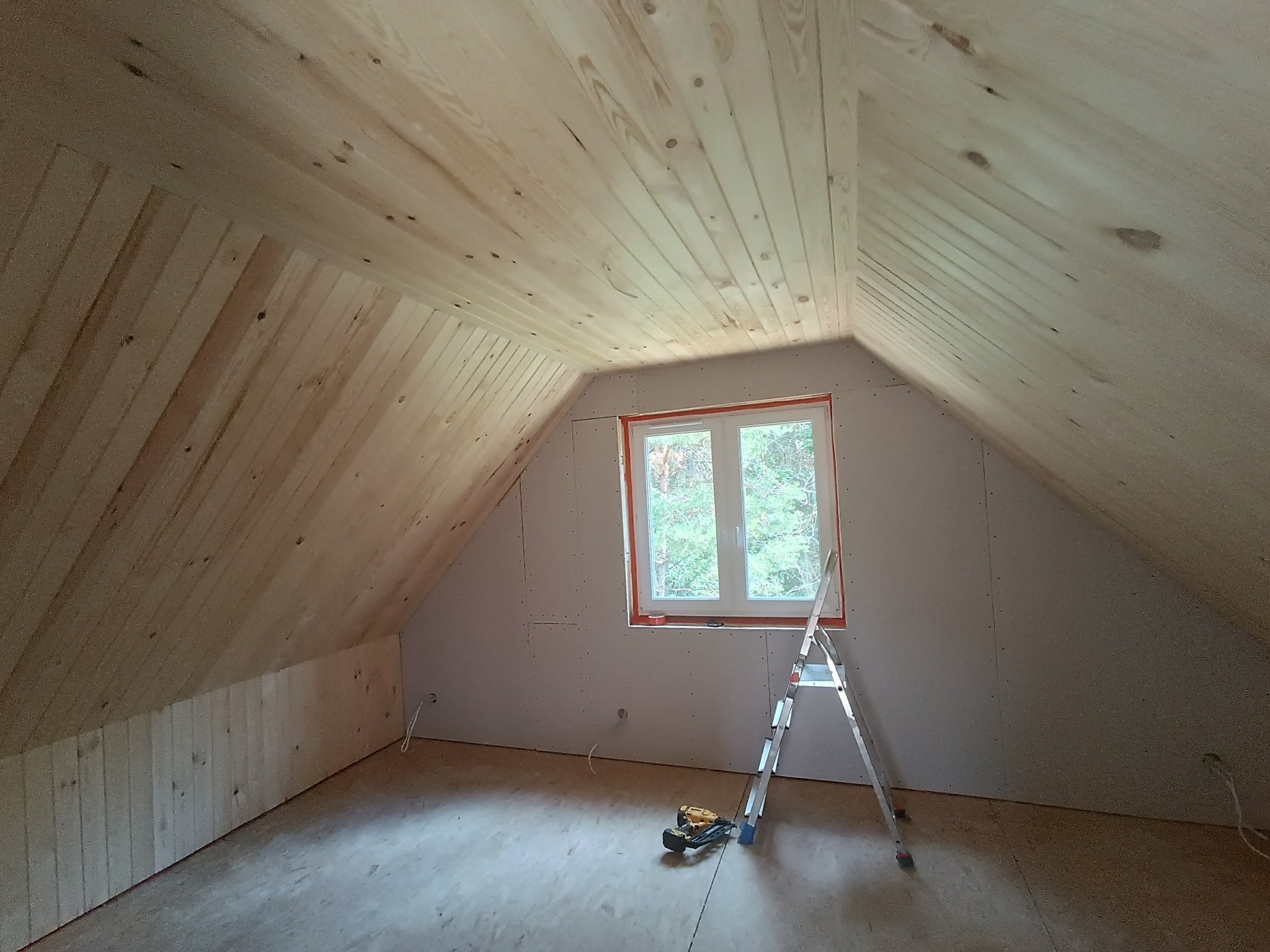 Poddasze w trakcie wykańczania, widoczne drewniane panele na skośnym suficie, ściany z płyt gipsowo-kartonowych, okno, drabina i narzędzia budowlane na podłodze.