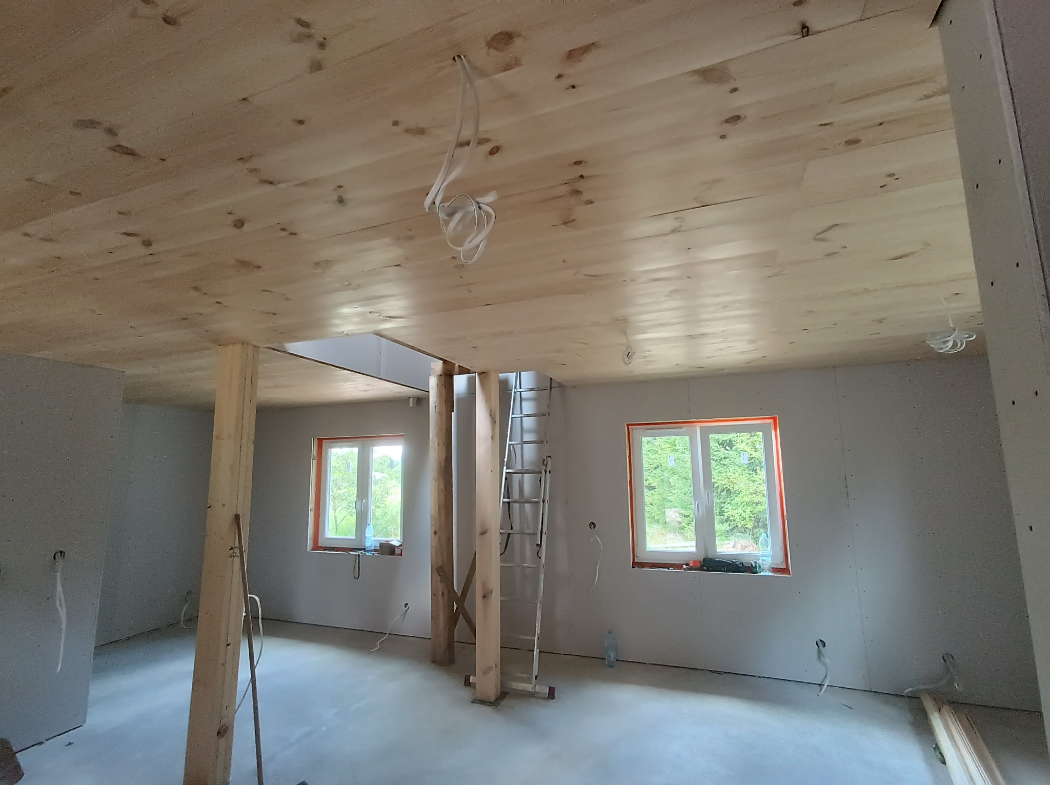 Wnętrze domu w trakcie budowy, widoczne drewniane belki stropowe, ściany w surowym stanie, okna z widokiem na zieleń, drabina oparta o belkę, oraz wystające kable elektryczne.