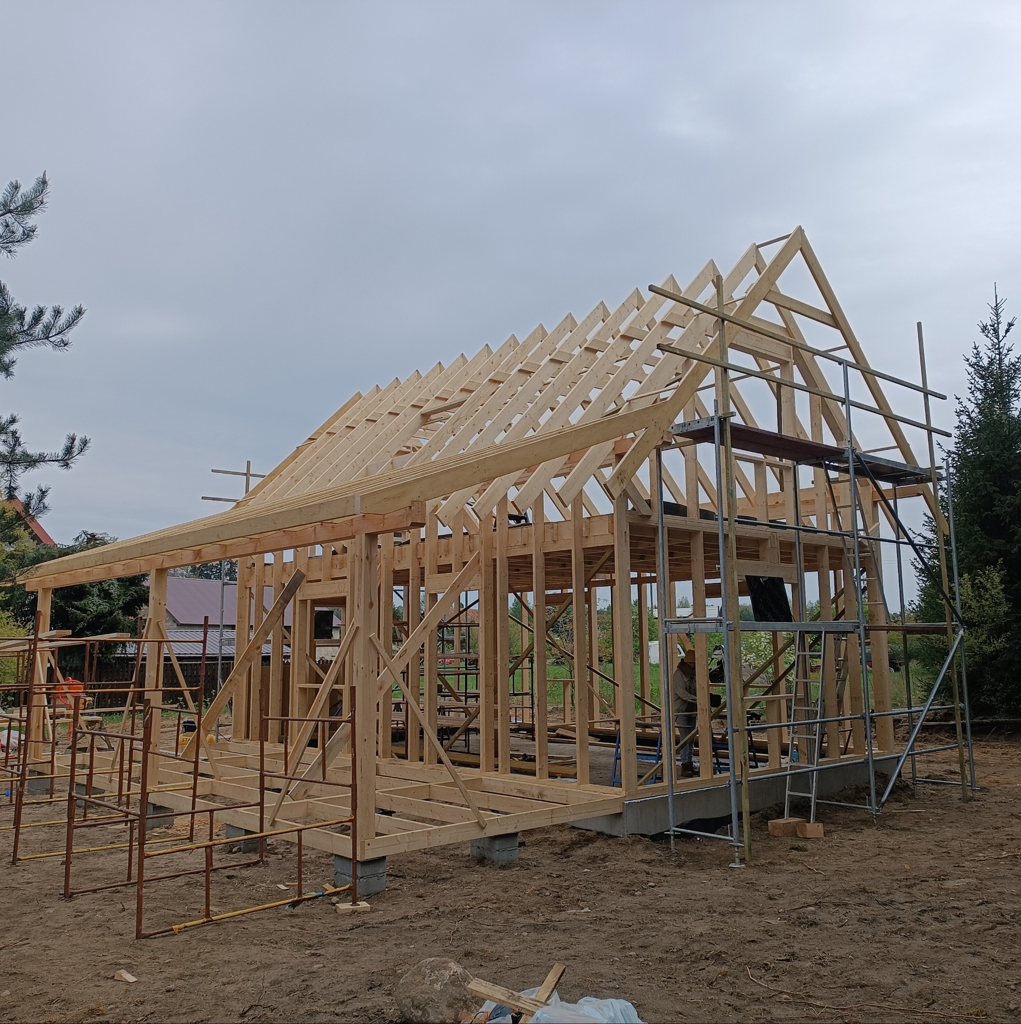 Konstrukcja szkieletowa domu w trakcie budowy, widoczne drewniane elementy ścian i dachu, rusztowania oraz fundamenty, na tle pochmurnego nieba.