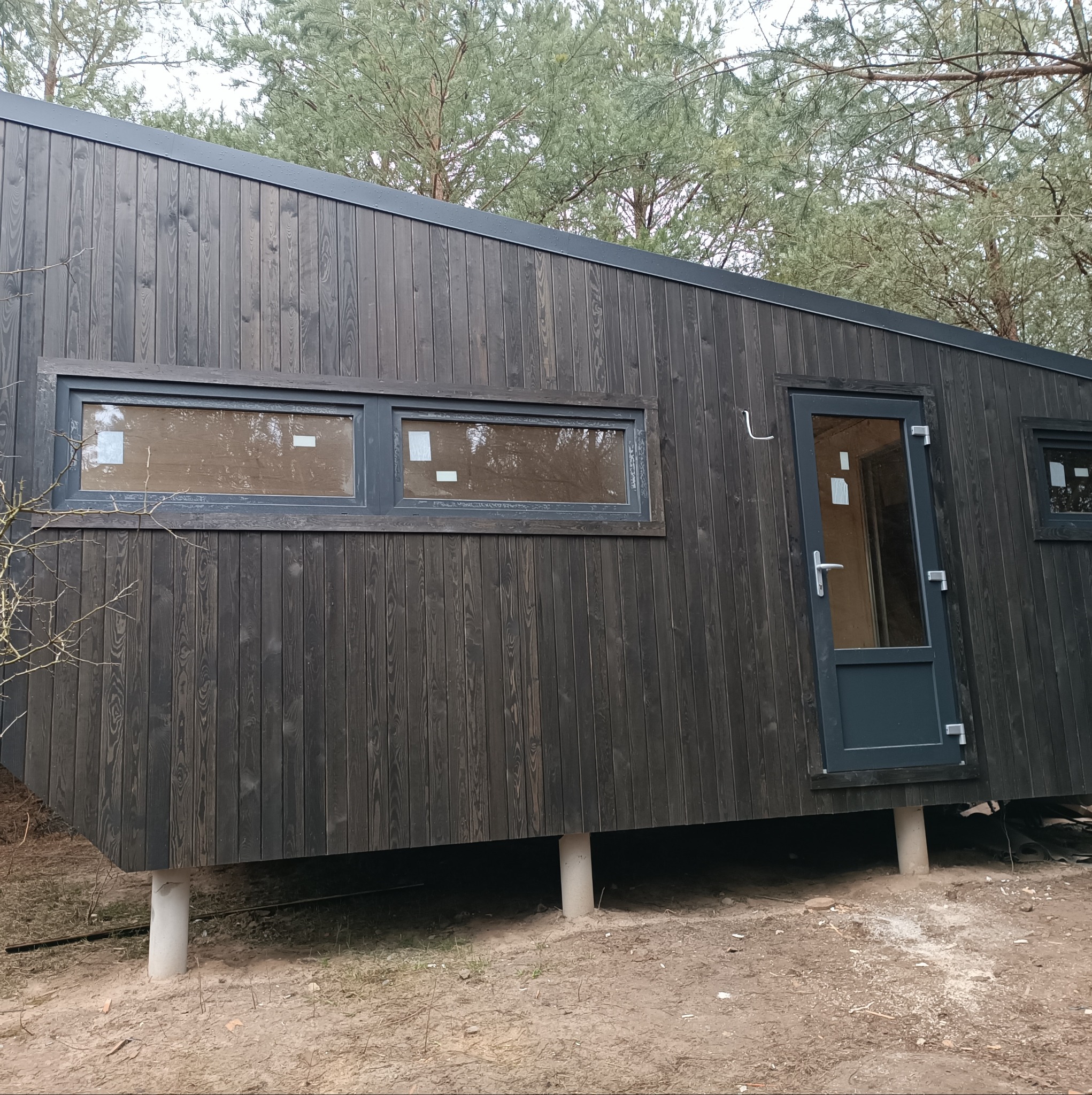 Ciemna, nowoczesna bryła domu z drewnianą elewacją, osadzona na betonowych słupach, z widocznymi oknami i drzwiami, otoczona drzewami.