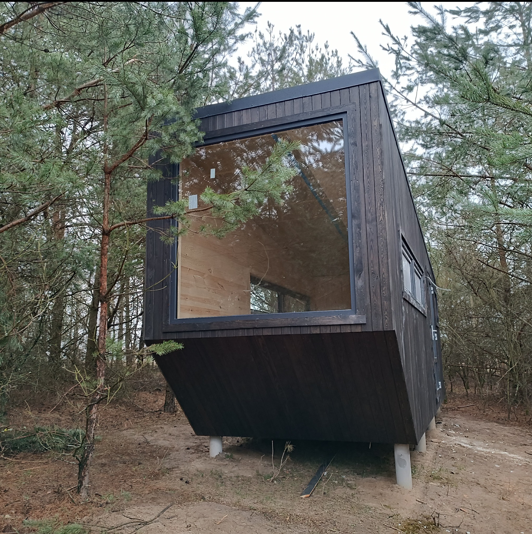 Czarny, nowoczesny budynek z dużym oknem, posadowiony na betonowych słupach, otoczony drzewami.