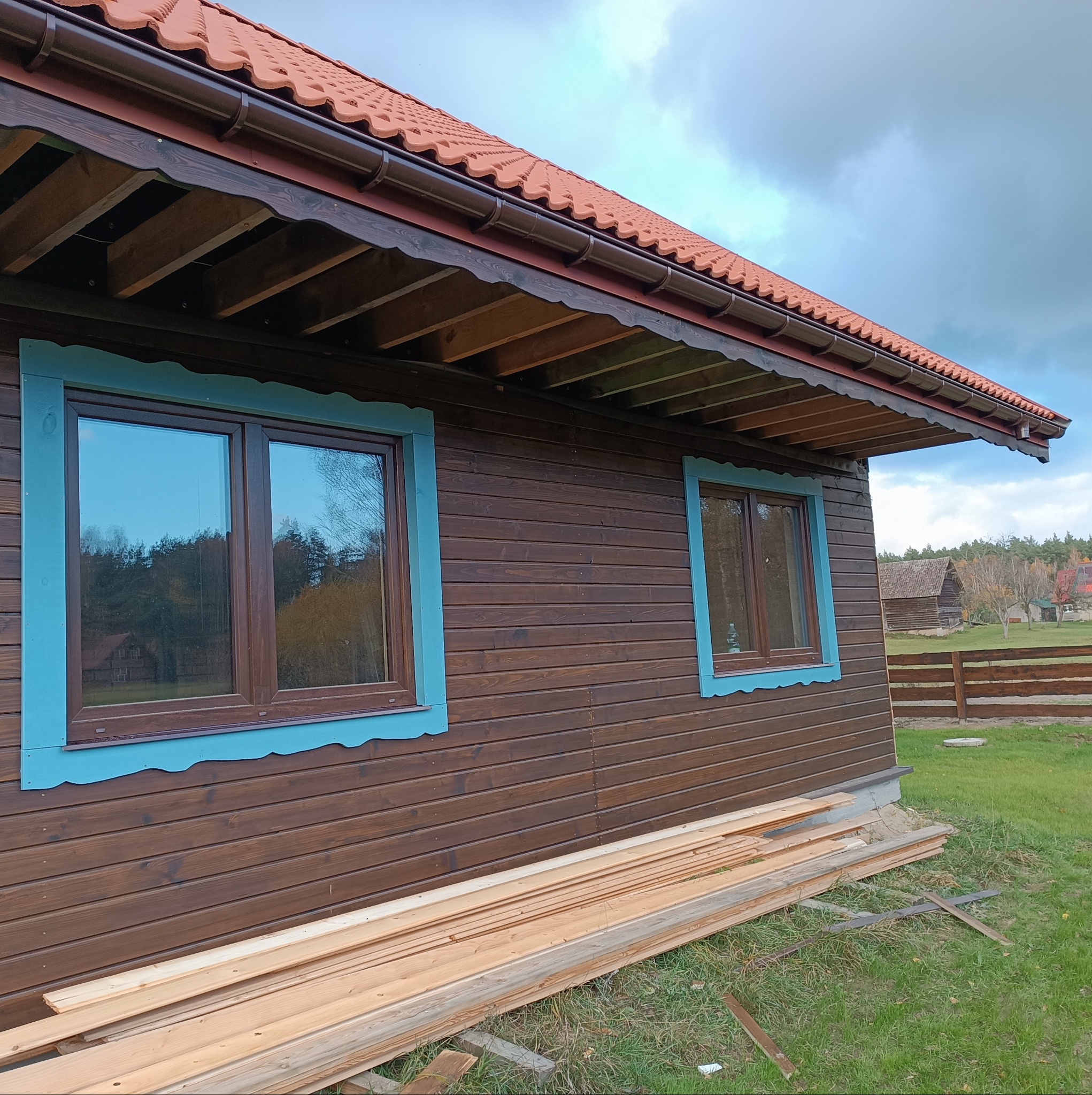 Ciemnobrązowy budynek z drewnianą elewacją i oknami z turkusowymi ramami, na tle zielonego trawnika i pochmurnego nieba, z ułożonymi deskami u podstawy ściany.