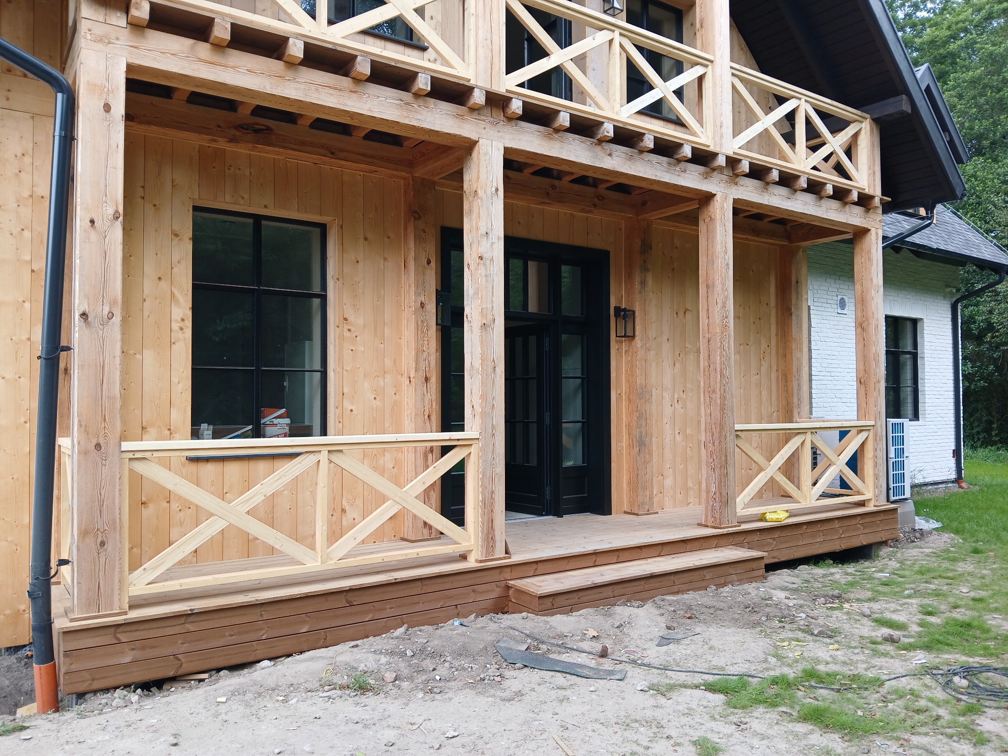 Surowy dom szkieletowy z drewnianą werandą i czarną stolarką okienną. Widoczne detale konstrukcyjne, naturalne drewno i fragment białej elewacji.