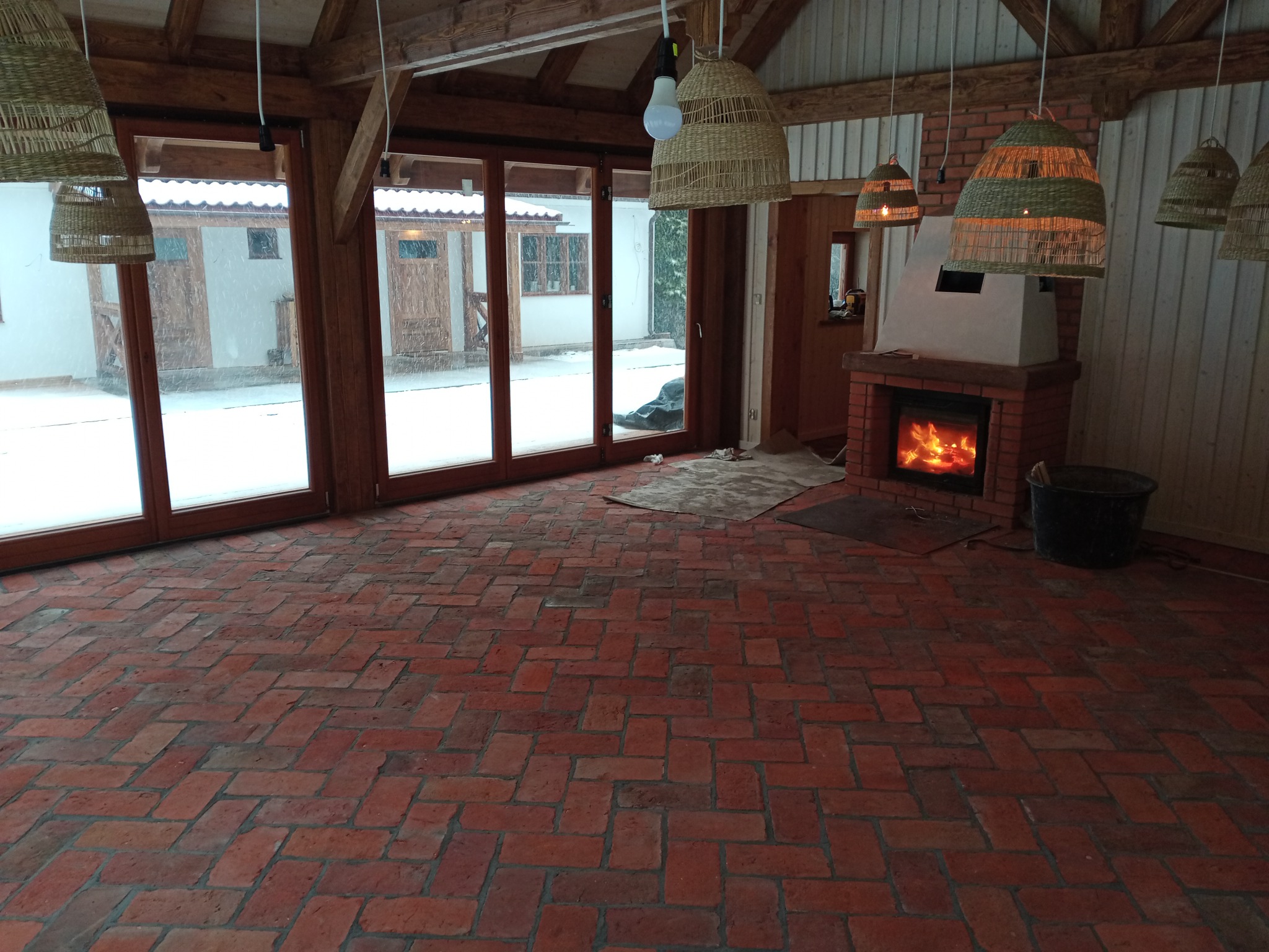 Jasne wnętrze z ceglaną podłogą ułożoną w jodełkę. Duże okna z widokiem na zimowy krajobraz. Kominek z paleniskiem i wiszące lampy z abażurami z naturalnych materiałów.