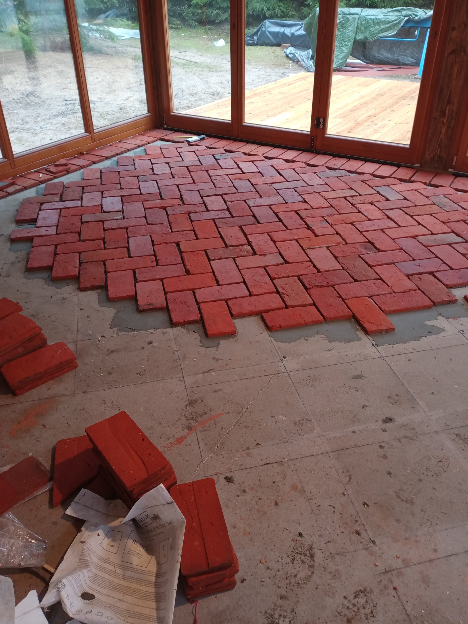 Układanie czerwonej cegły klinkierowej na podłodze w przeszklonym pomieszczeniu. Widoczne częściowo ułożone cegły i zapas materiału obok. Praca w toku.