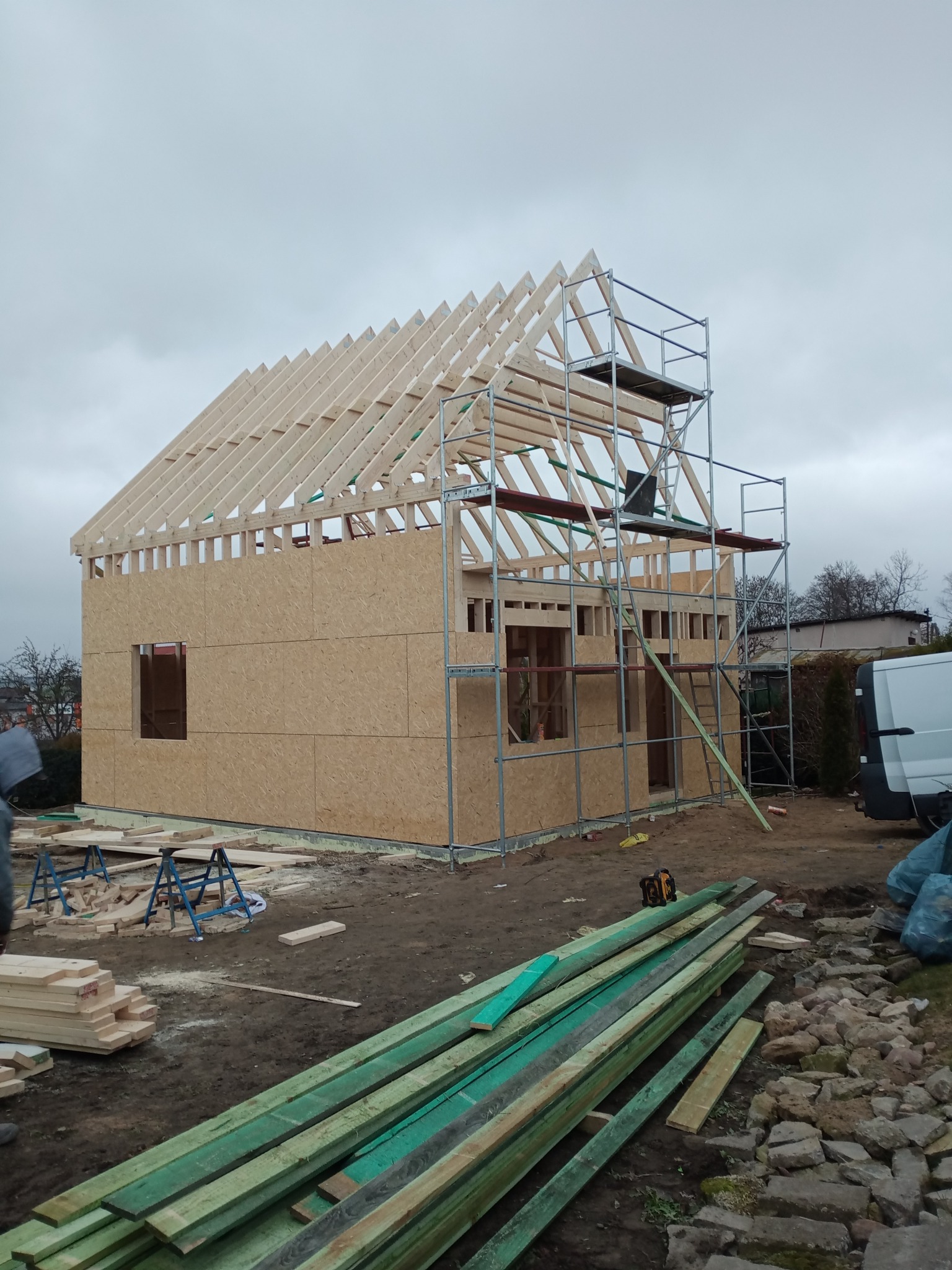 Wznoszenie domu szkieletowego: widok na konstrukcję z płyt OSB, rusztowania i drewniane elementy dachu w trakcie montażu. Na pierwszym planie stos drewna.