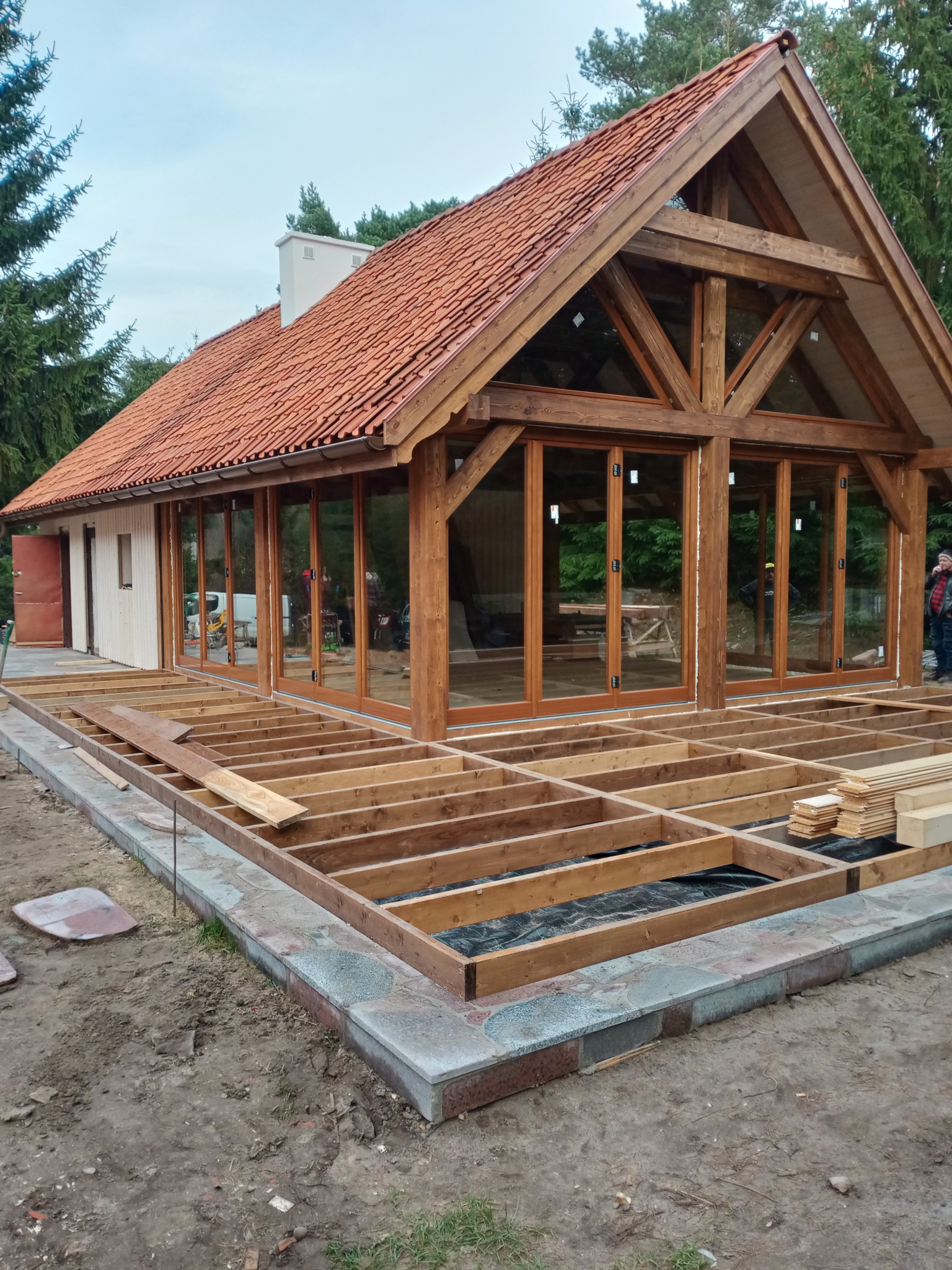 Budowa drewnianego domu z tarasem w trakcie realizacji: widoczne fundamenty tarasu, szkielet domu z dużymi oknami i czerwonym dachem. Etap budowy.