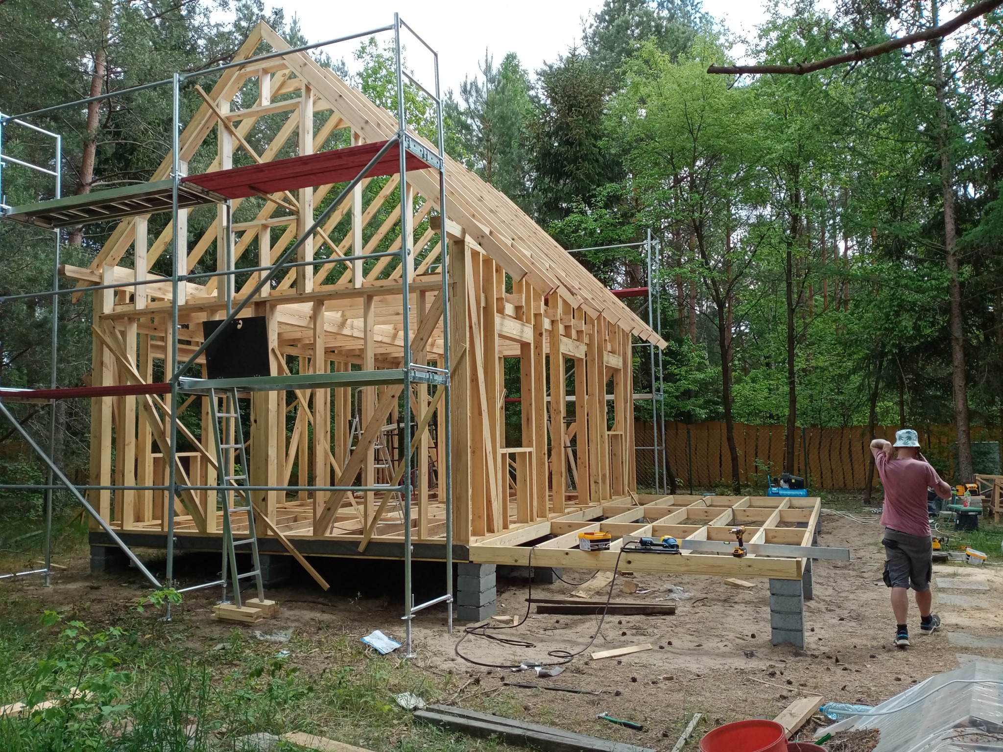 Konstrukcja szkieletowa domu w trakcie budowy, widoczne rusztowania i narzędzia na drewnianym fundamencie, otoczenie leśne, osoba w tle.