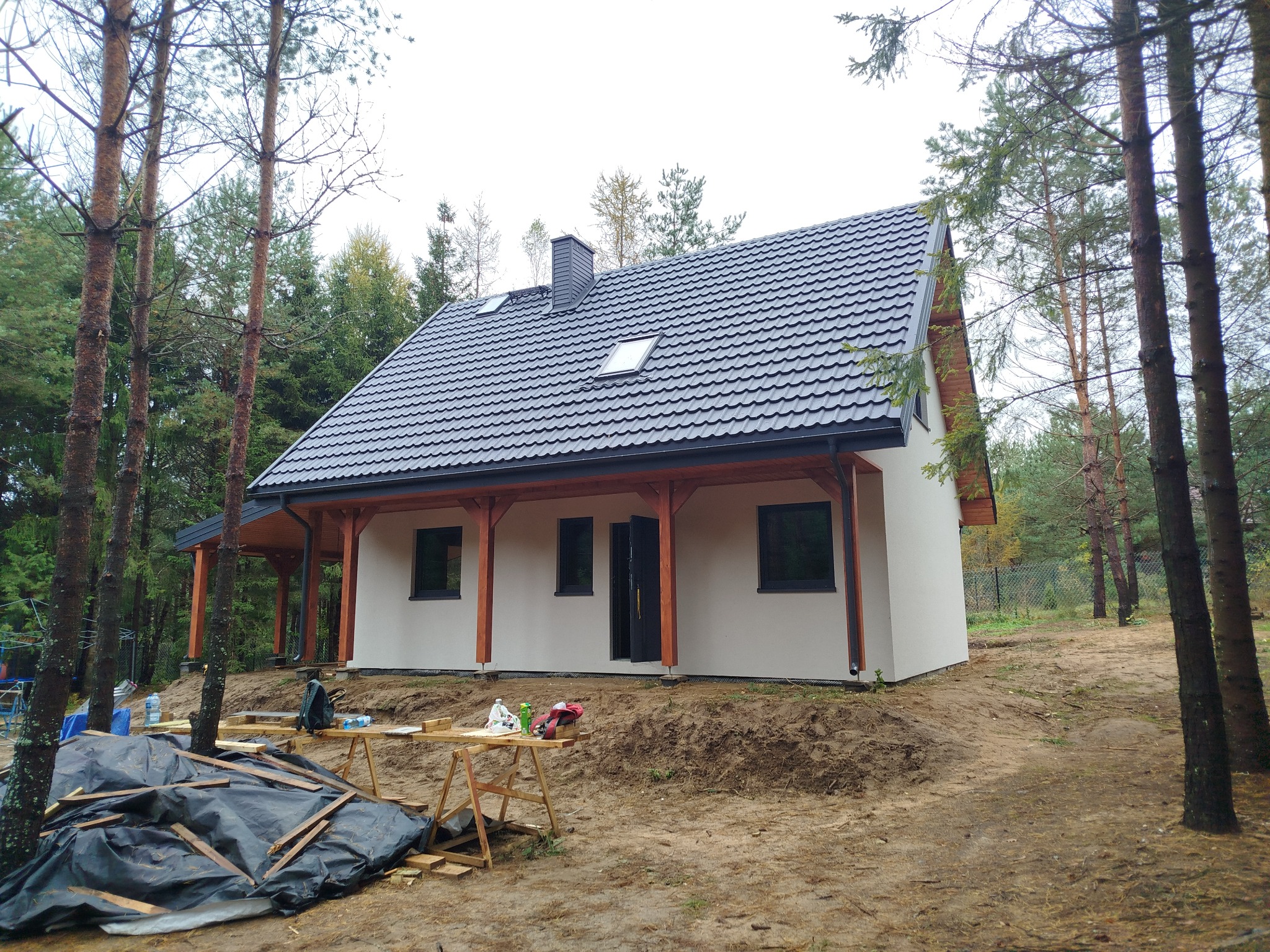 Nowoczesny dom z ciemną dachówką i drewnianą konstrukcją werandy, otoczony sosnowym lasem. Widoczne narzędzia sugerują trwające prace wykończeniowe.