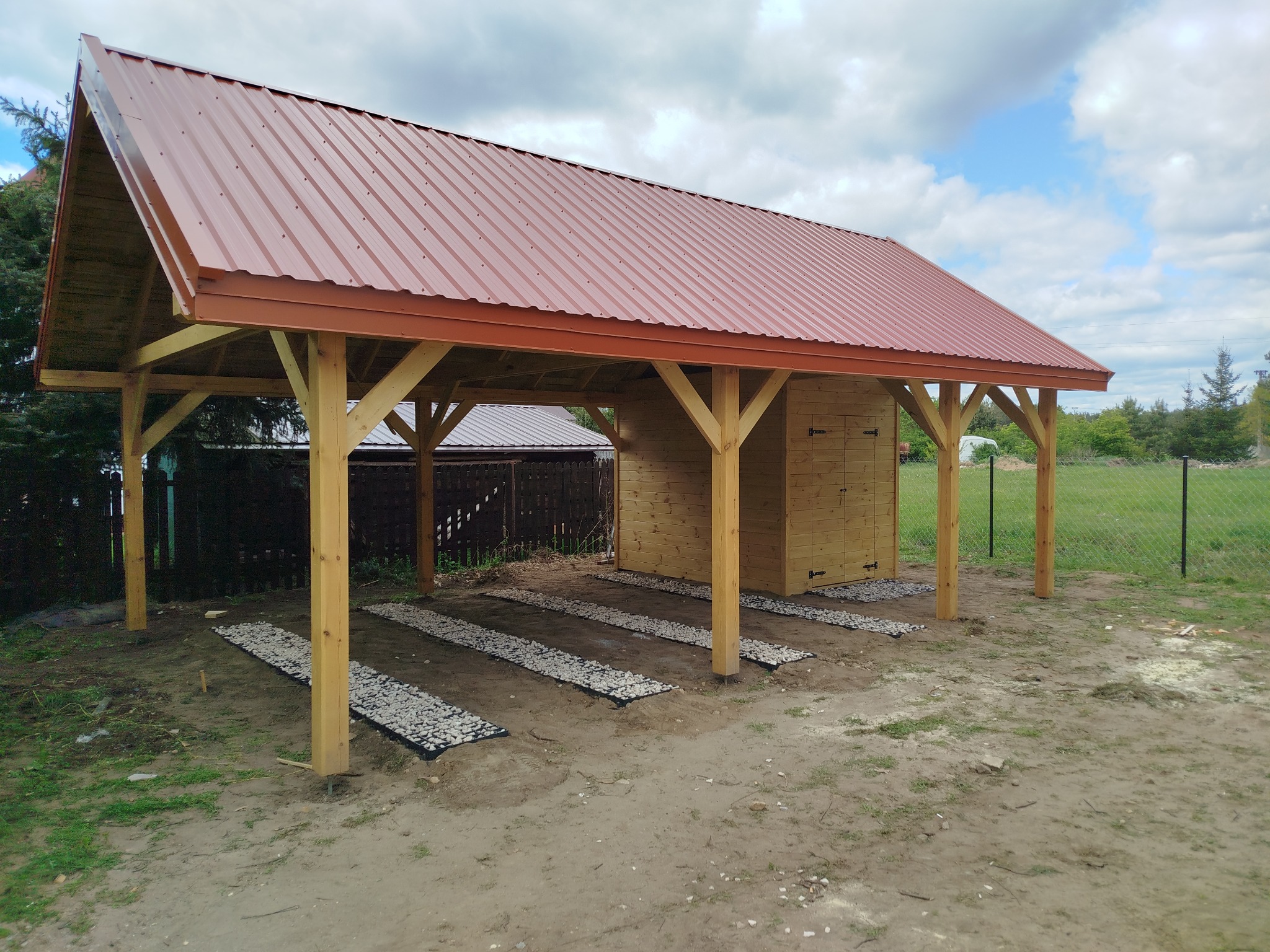 Drewniana altana z czerwonym dachem blaszanym w trakcie budowy. Widoczne przygotowane podłoże z kamieni. W tle zieleń i błękitne niebo z chmurami.