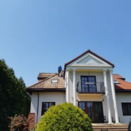Pracownik w niebieskim kombinezonie myje dach z czerwonej dachówki na dwupiętrowym domu z kolumnami, widoczne narzędzia i drabina oparta o ścianę