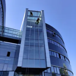 Trzech alpinistów na linach zabezpieczających czyści szklaną fasadę nowoczesnego biurowca w słoneczny dzień, widok od dołu.